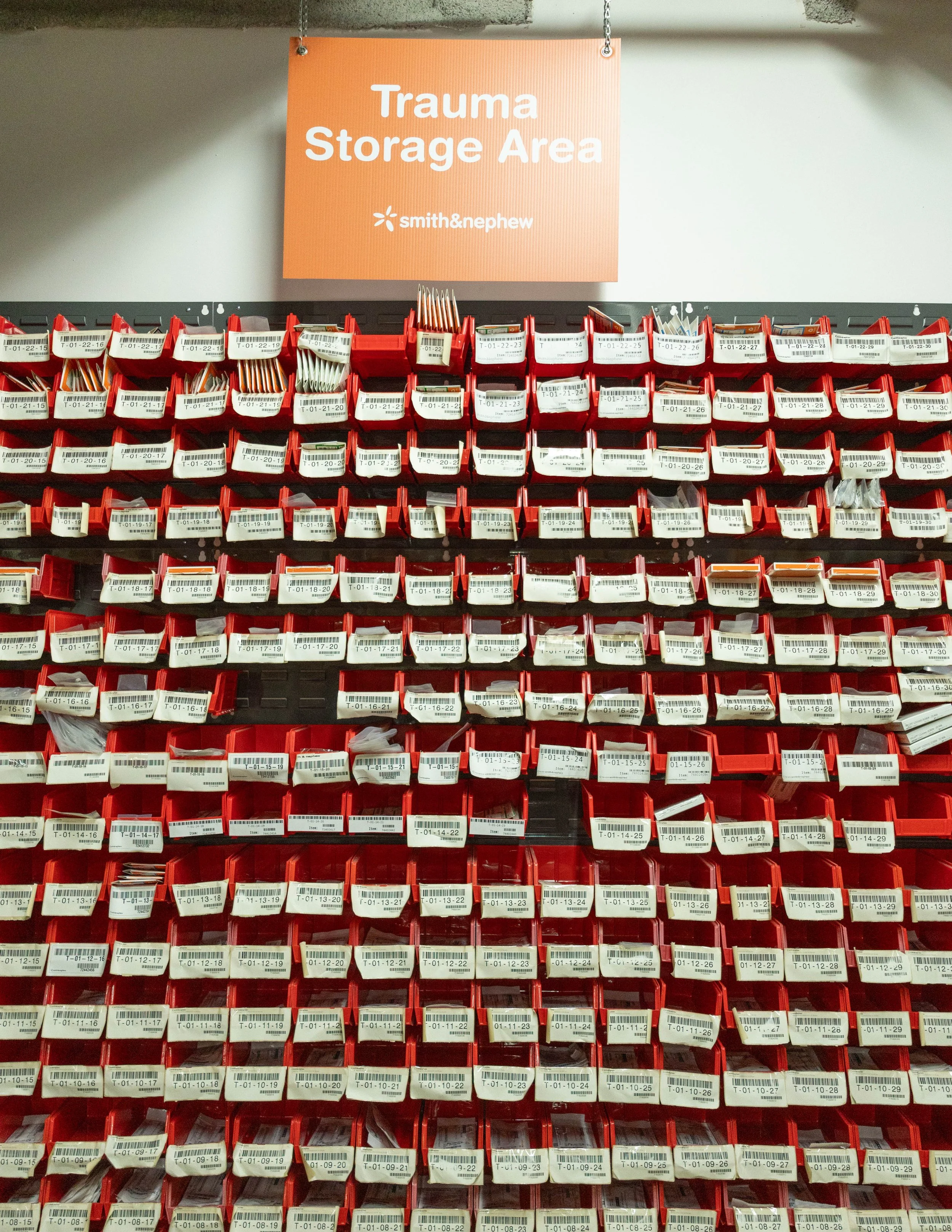Red storage bins with medical supplies organized on a wall, labeled with codes, in a trauma storage area.