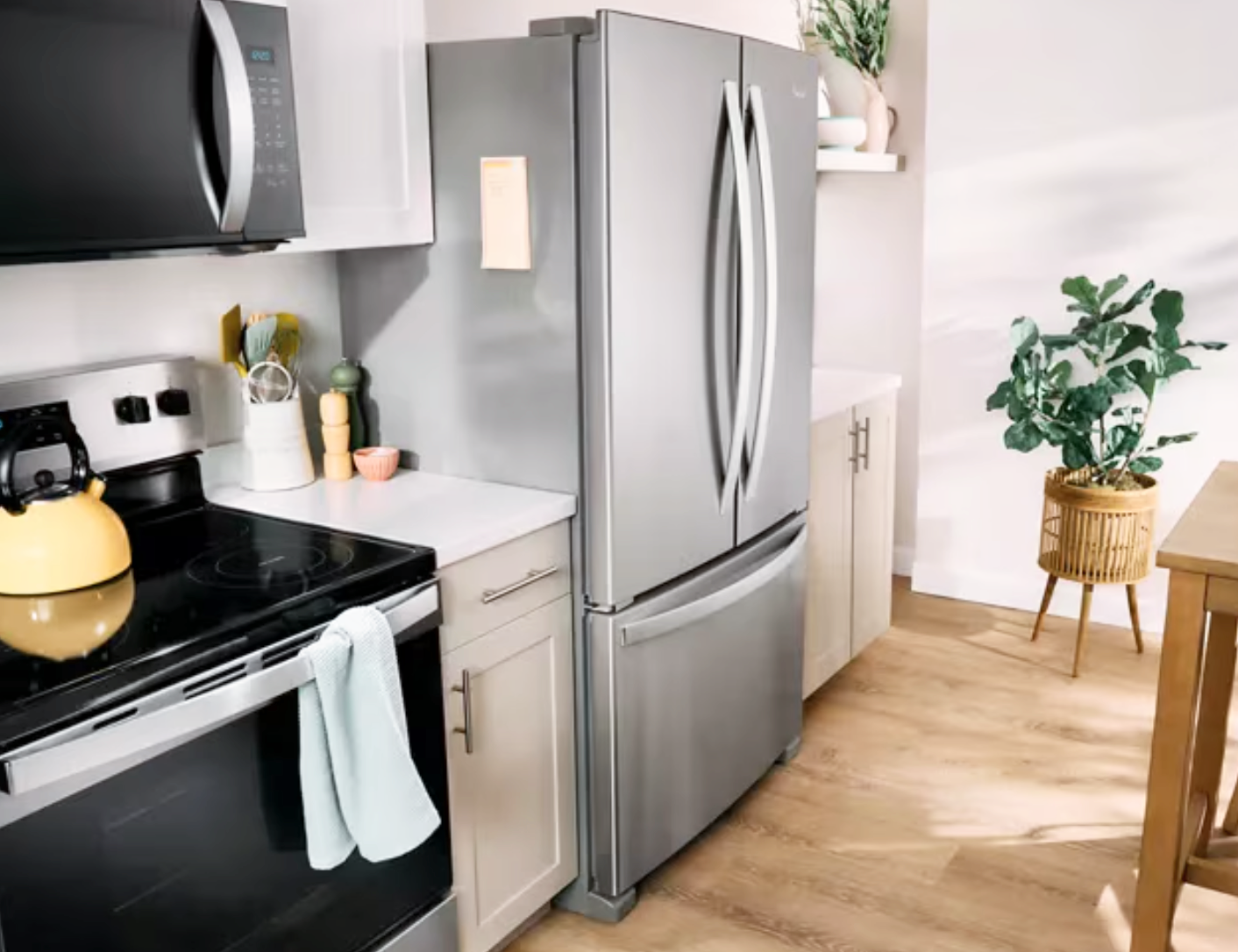 Modern kitchen with stainless steel refrigerator, stove with a tea kettle, white cabinets, and a countertop with kitchen utensils and a small potted plant. Wooden floor, open shelves, and a large leafy plant in a wicker basket pot near a wooden table.