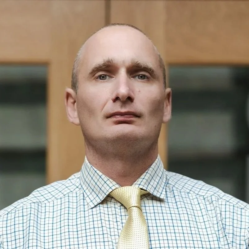A smiling man in a suit with a striped tie, white shirt, and a dark blazer, standing indoors.
