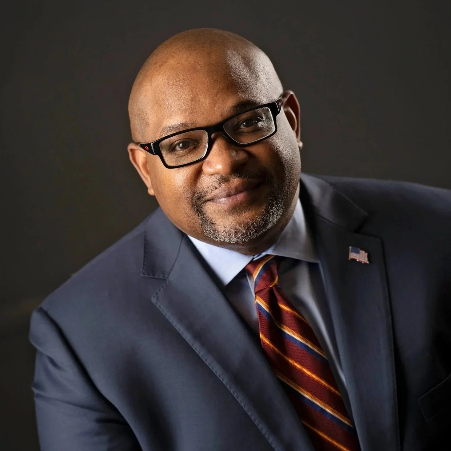Close-up portrait of a man in a suit with an American flag pin, wearing glasses, smiling against a dark background.