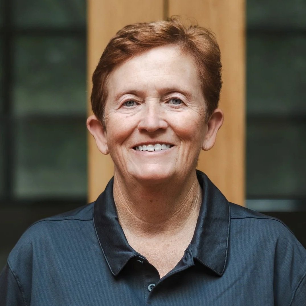 A woman with short red hair smiling, wearing a blue polo shirt with 'Nazareth Prep' logo, standing against a dark gray background.