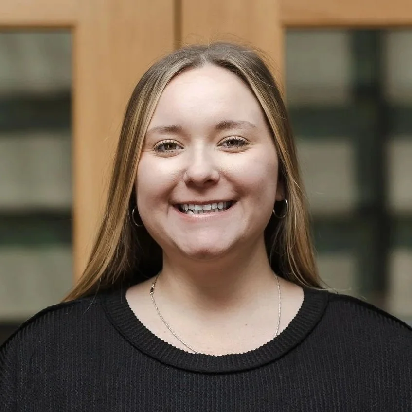 A young woman with long blonde hair, wearing a black lace top, smiling against a plain light-colored wall.