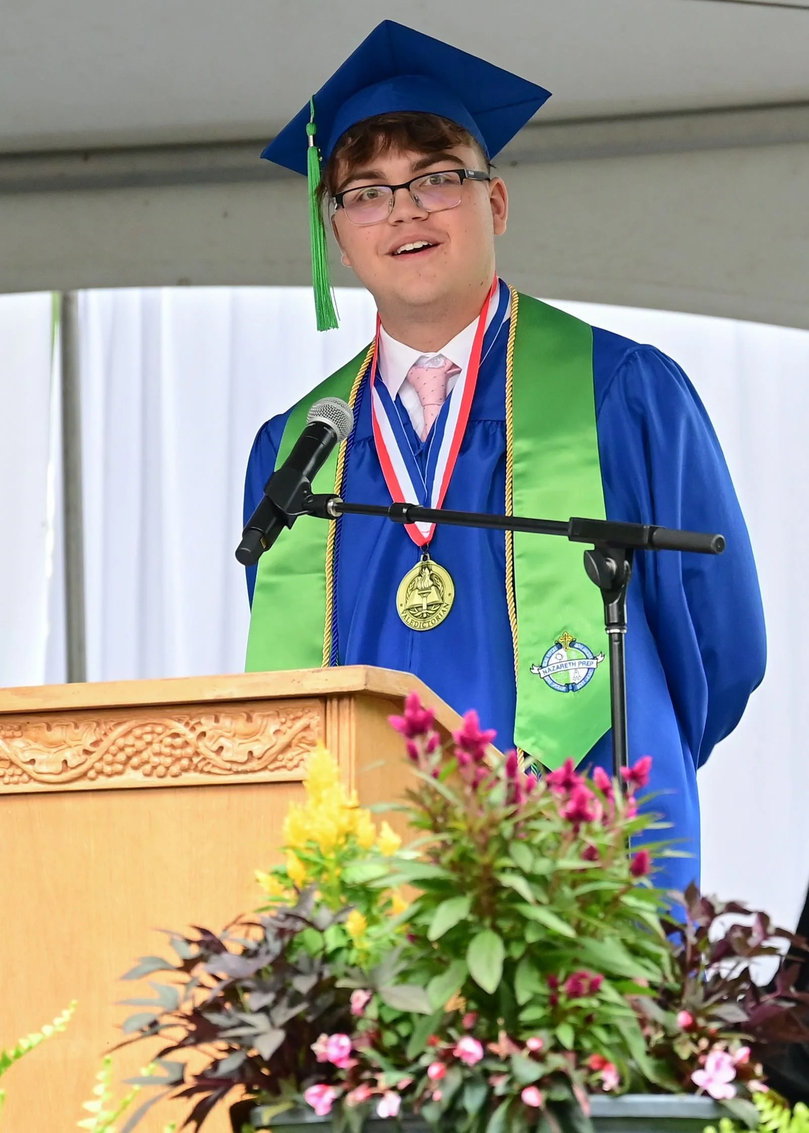A photo of a young man wearing blue and green graduation robe and cap speaking at a podium