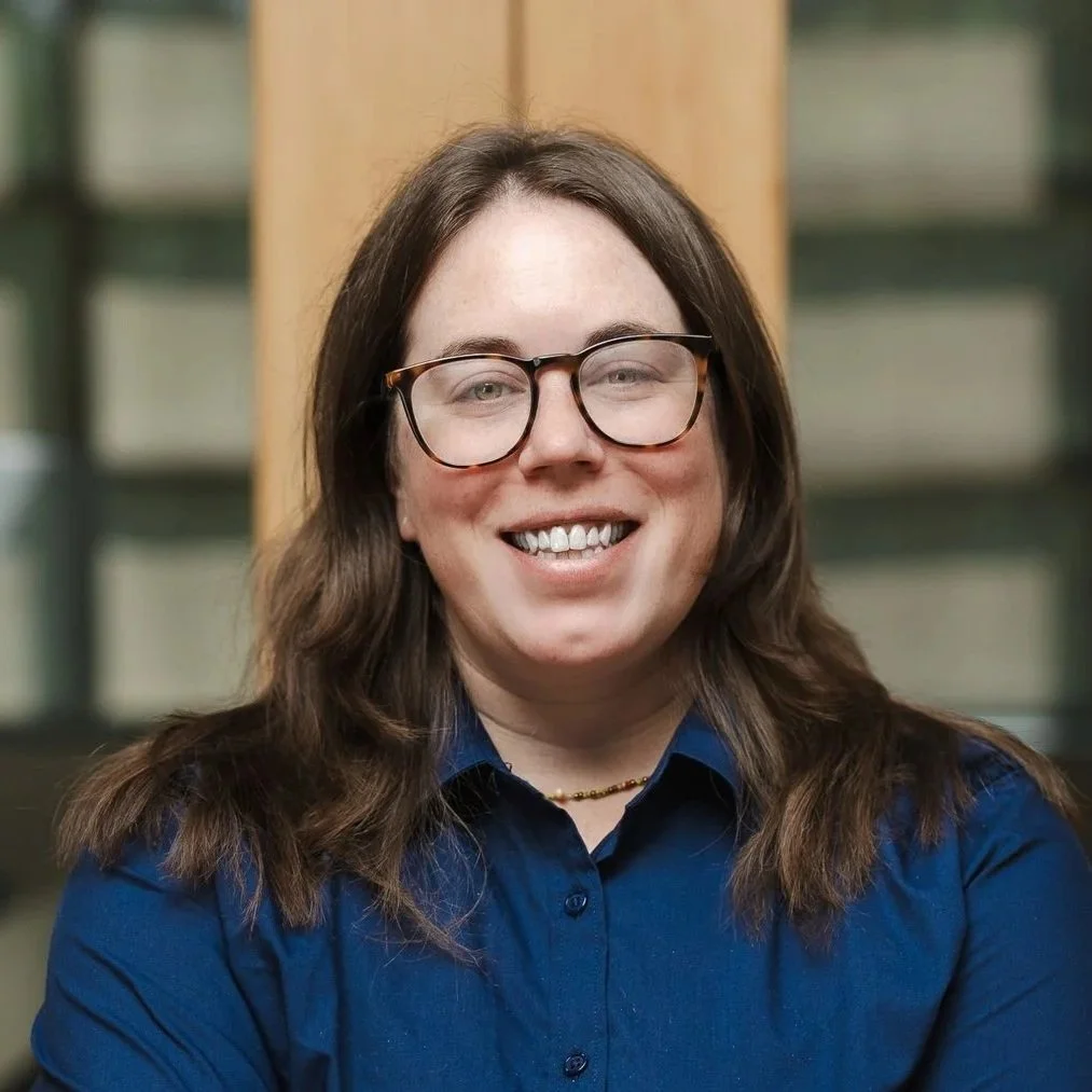 A woman with long brown hair, glasses, and a striped shirt smiling at the camera.