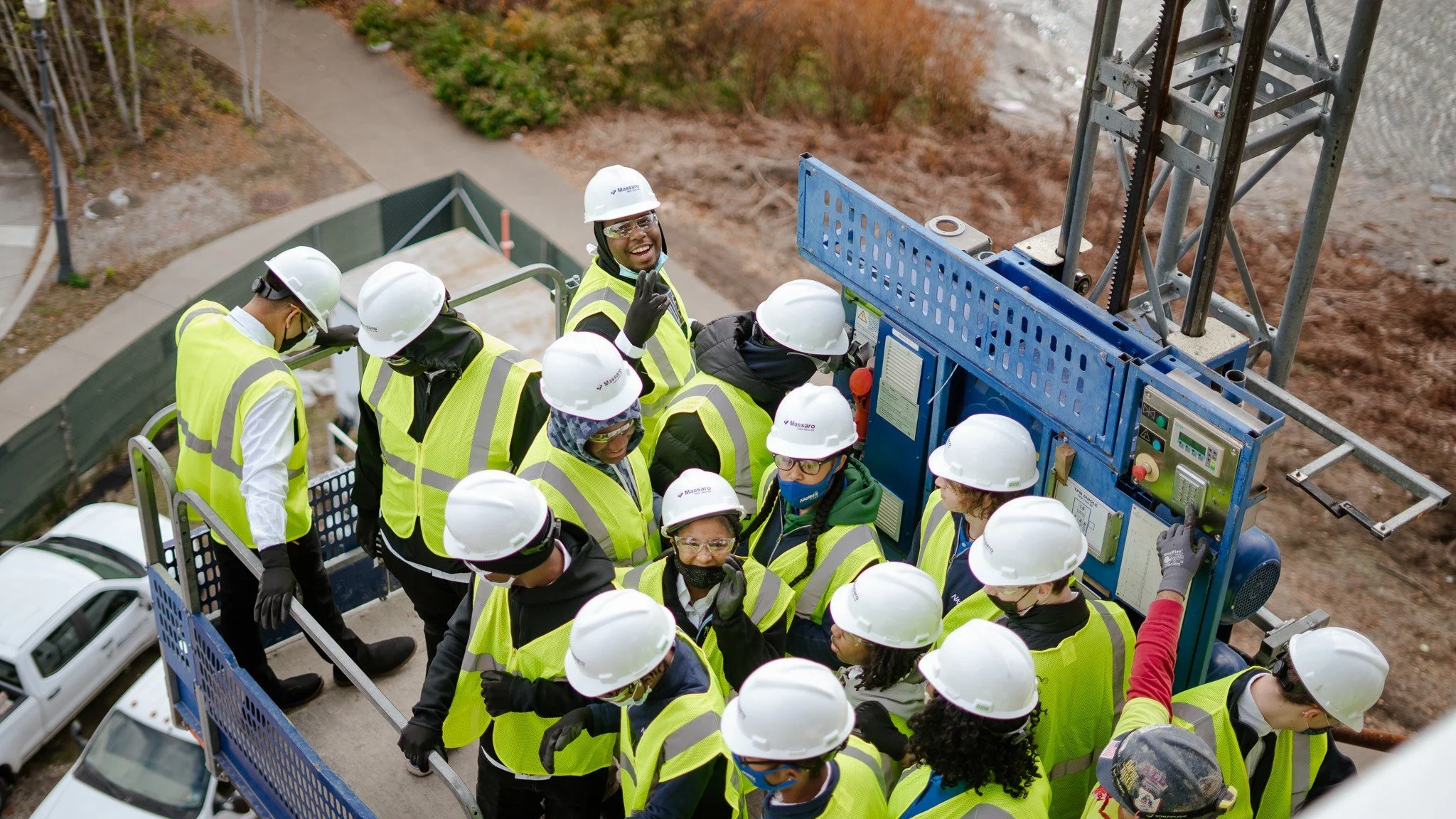 A group of students wearing hard hats, safety vests, and gloves gathered around a large blue machinery or generator on a construction site, with some students appearing to listen while others look at the machinery.