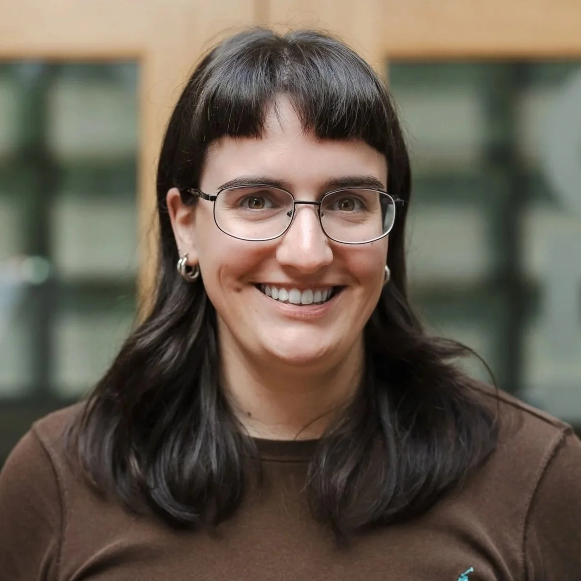 A woman with short dark hair, glasses, earrings, and a red and black patterned top smiling at the camera.