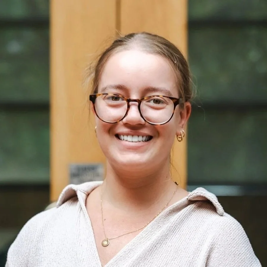 A woman with long blond hair, glasses, and gold hoop earrings smiling at the camera, wearing a pink striped top.