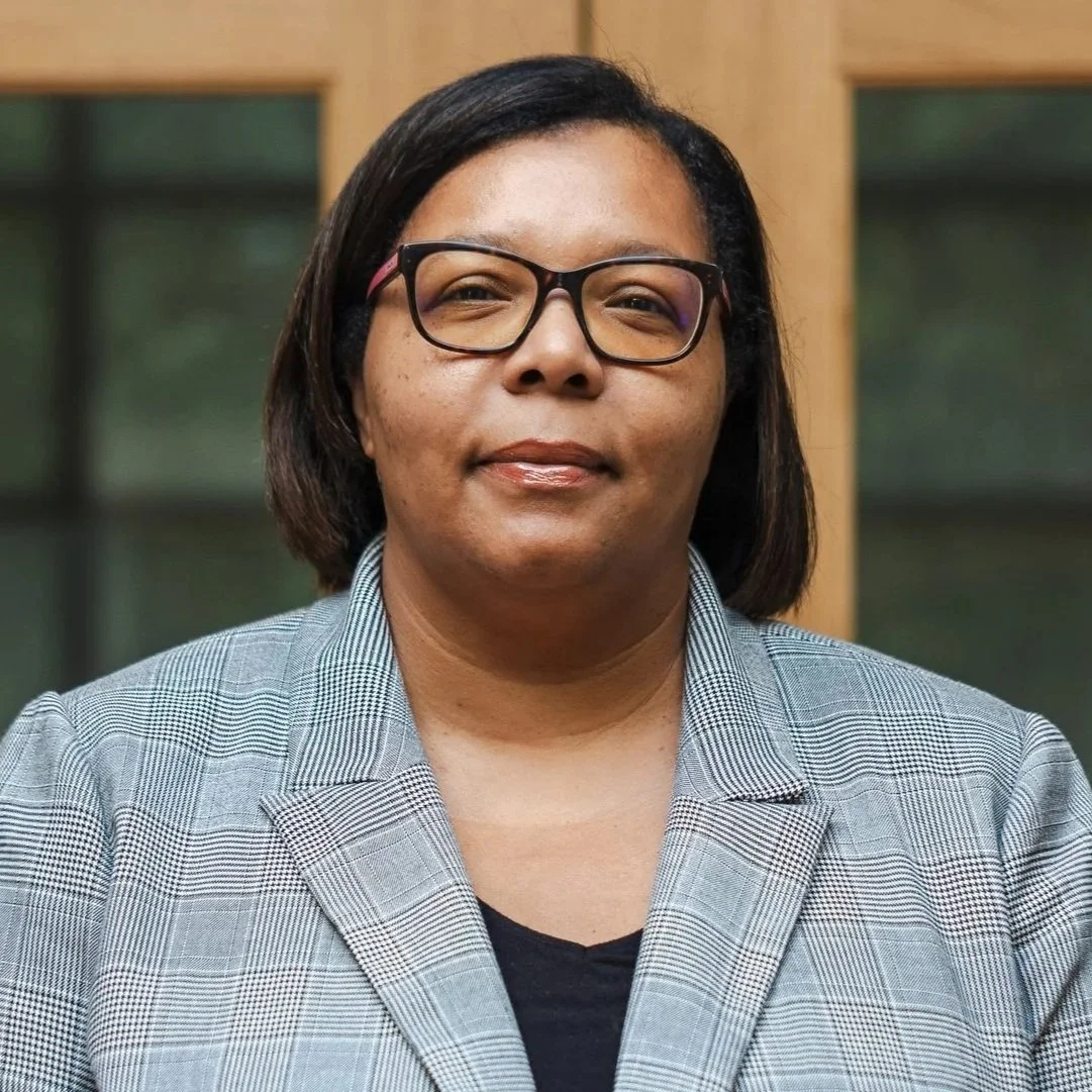 A woman with short hair, glasses, and a patterned top standing indoors near a window with sunlight, smiling.