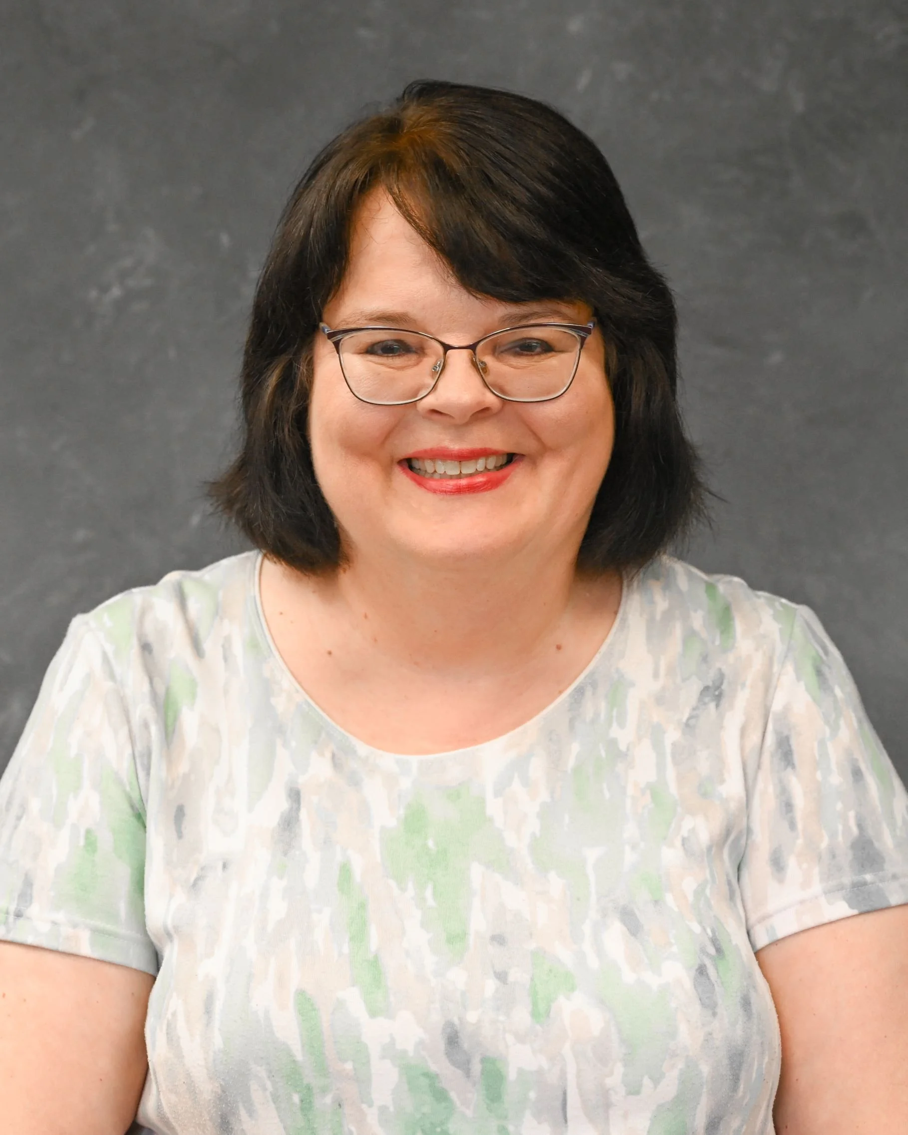 A smiling woman with glasses and dark shoulder-length hair, wearing a light-colored patterned blouse, sitting in front of a dark grey background.