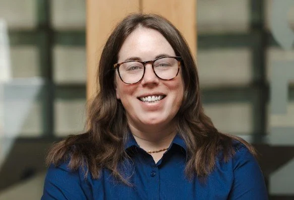 A woman with long brown hair, glasses, and a striped shirt smiling at the camera.