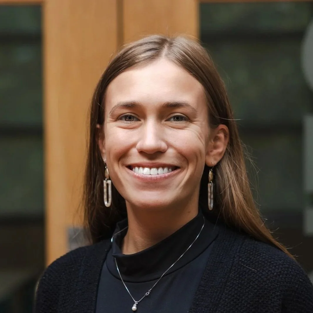 Smiling young woman with long hair, wearing earrings and a white top, standing indoors with a blurred background.