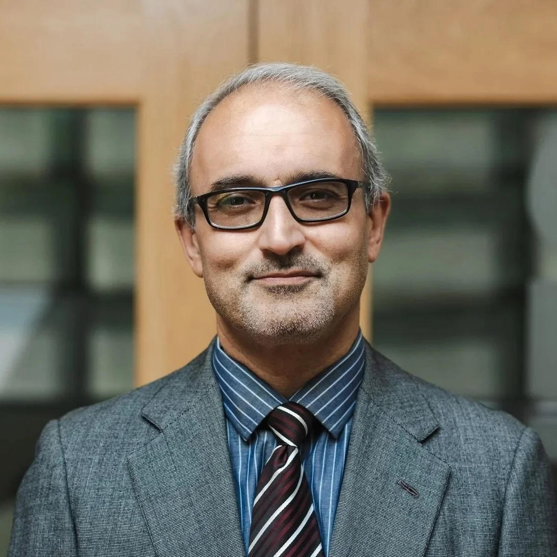 A gray-haired man with glasses, wearing a jacket and tie, smiling at the camera.