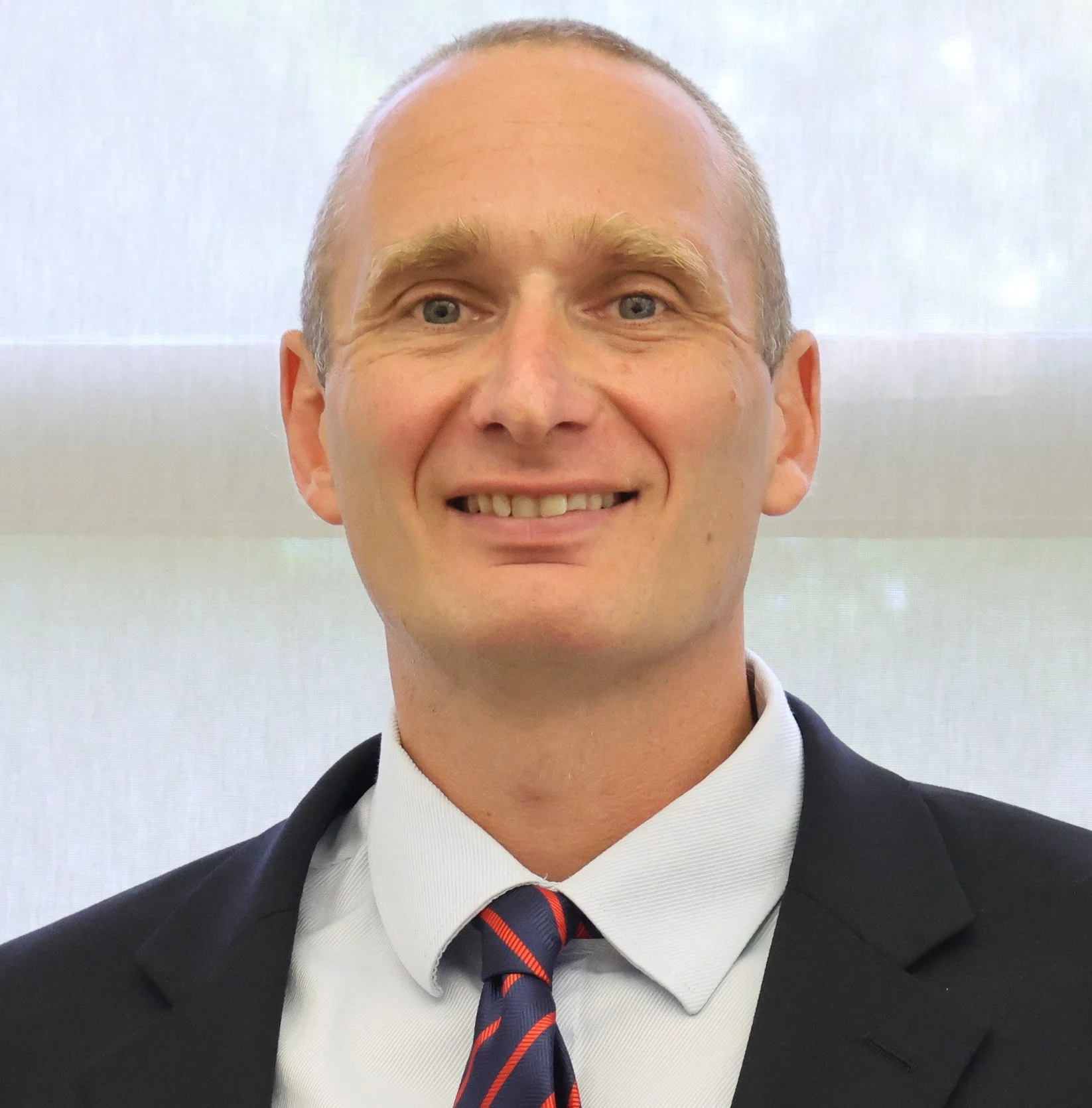 A smiling man in a suit with a striped tie, white shirt, and a dark blazer, standing indoors.