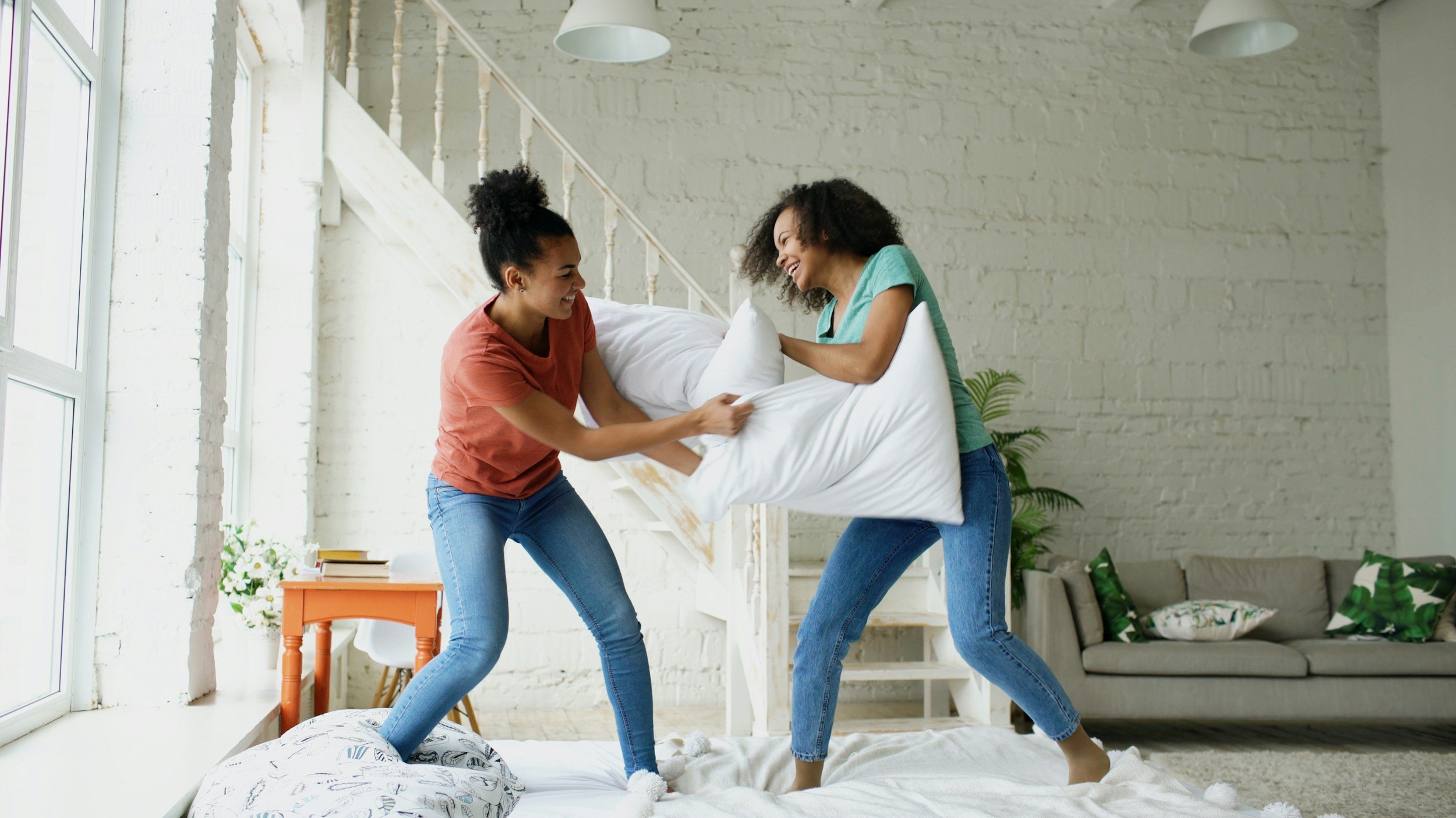 Two women are playful fighting with pillows on a bed in a bright, modern living room.
