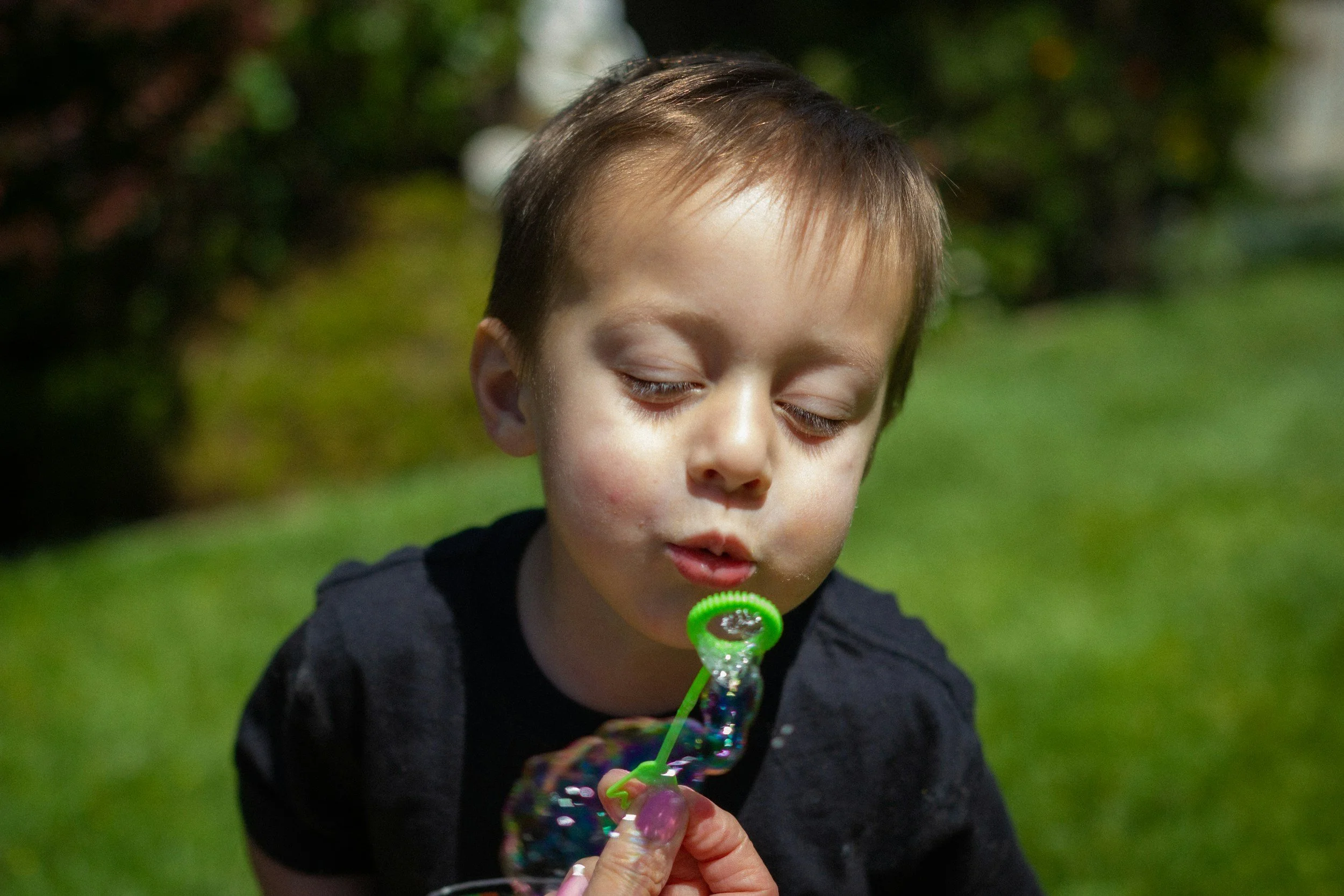 A young boy with closed eyes blowing bubbles outdoors on a grassy area with blurred green foliage in the background.