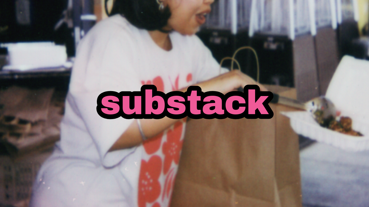 A girl wearing a white t-shirt with orange patterns, standing at a counter with a brown paper bag, looking at a food box with food inside in a setting that appears to be a cafeteria or food court.