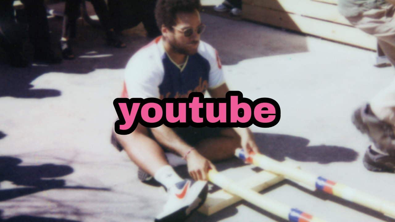 Young man wearing sunglasses and a sports jersey kneeling on the ground and holding two PVC pipes attached to a wooden frame, outdoors with other people around.