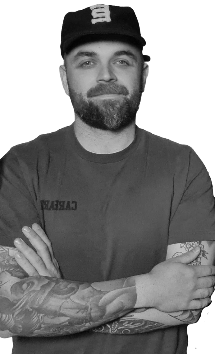 A man with a beard wearing a black baseball cap and a t-shirt, standing with arms crossed, displaying colorful tattoos on his left arm.