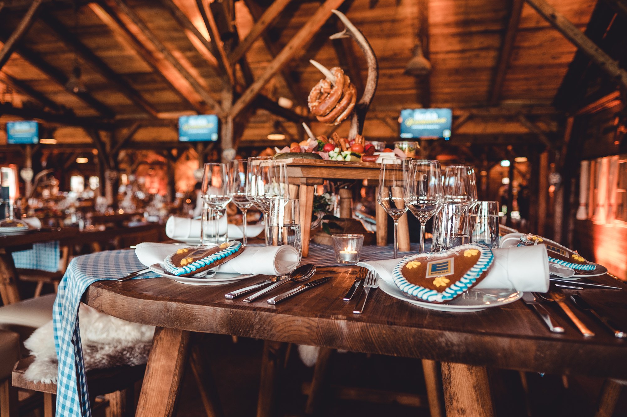Rustikale Eventlocation mit festlich dekorierten Holztischen, Gläsern und bayerischen Details. Komplettpakete für Firmenfeier-Ausstattung im Oktoberfest- oder Hüttenstil mieten.