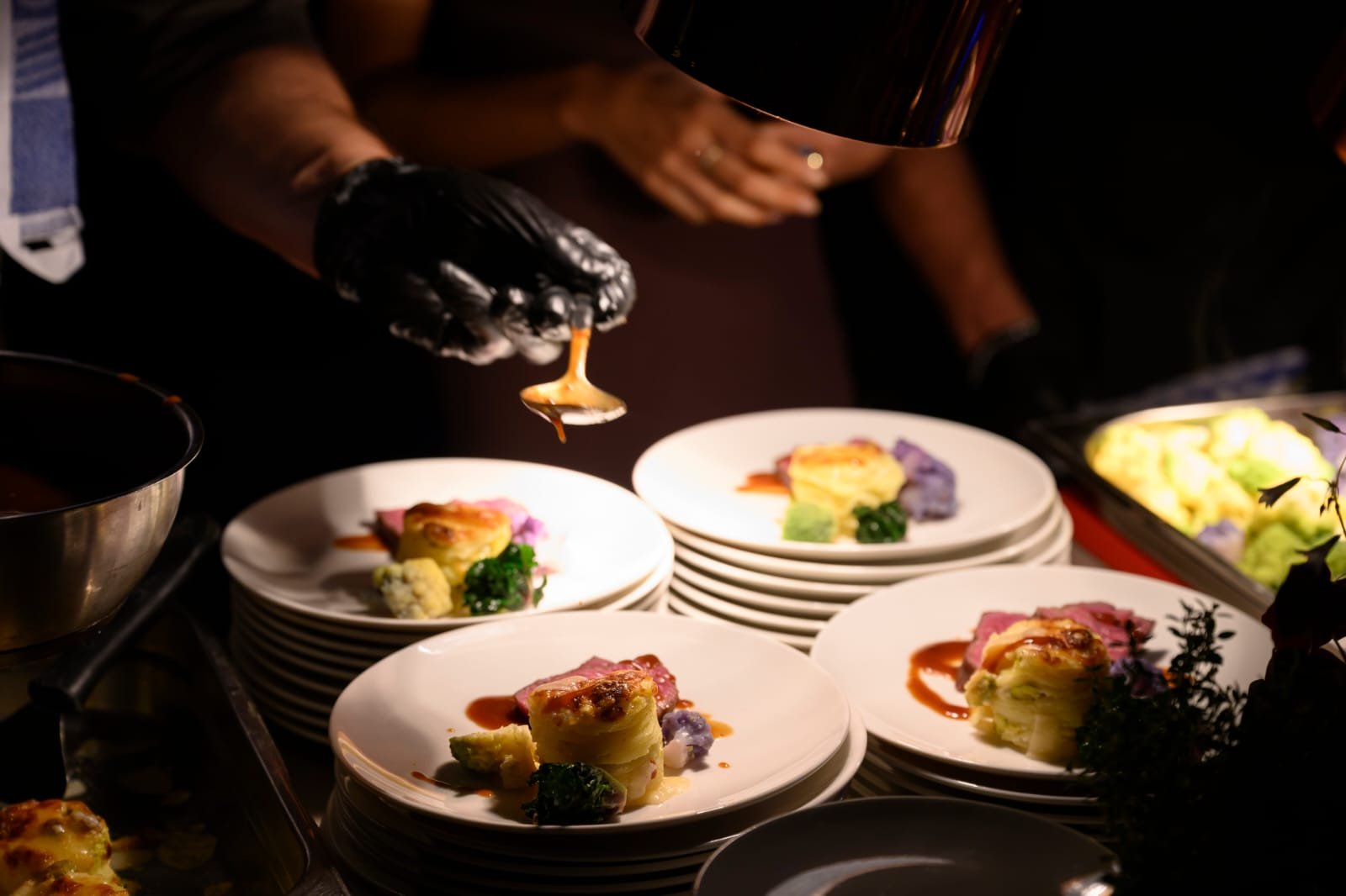 Koch serviert auf weißen Tellern mit Speisen, in gläsernem Löffel Soße, unter dunklem Licht auf einem Buffet.