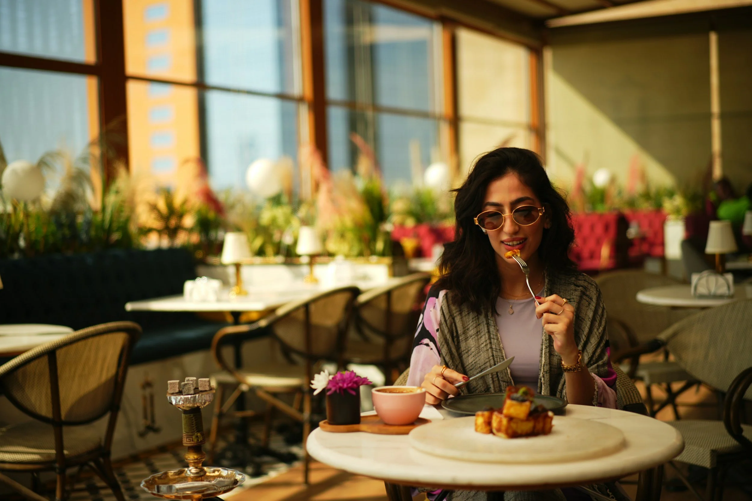 A woman with dark wavy hair, sunglasses, and a patterned shawl is sitting at a table in a bright, modern restaurant, eating a piece of cake. The table has a bowl of soup, a small plant, and a plate with toast. The background features large windows and colorful floral decorations.
