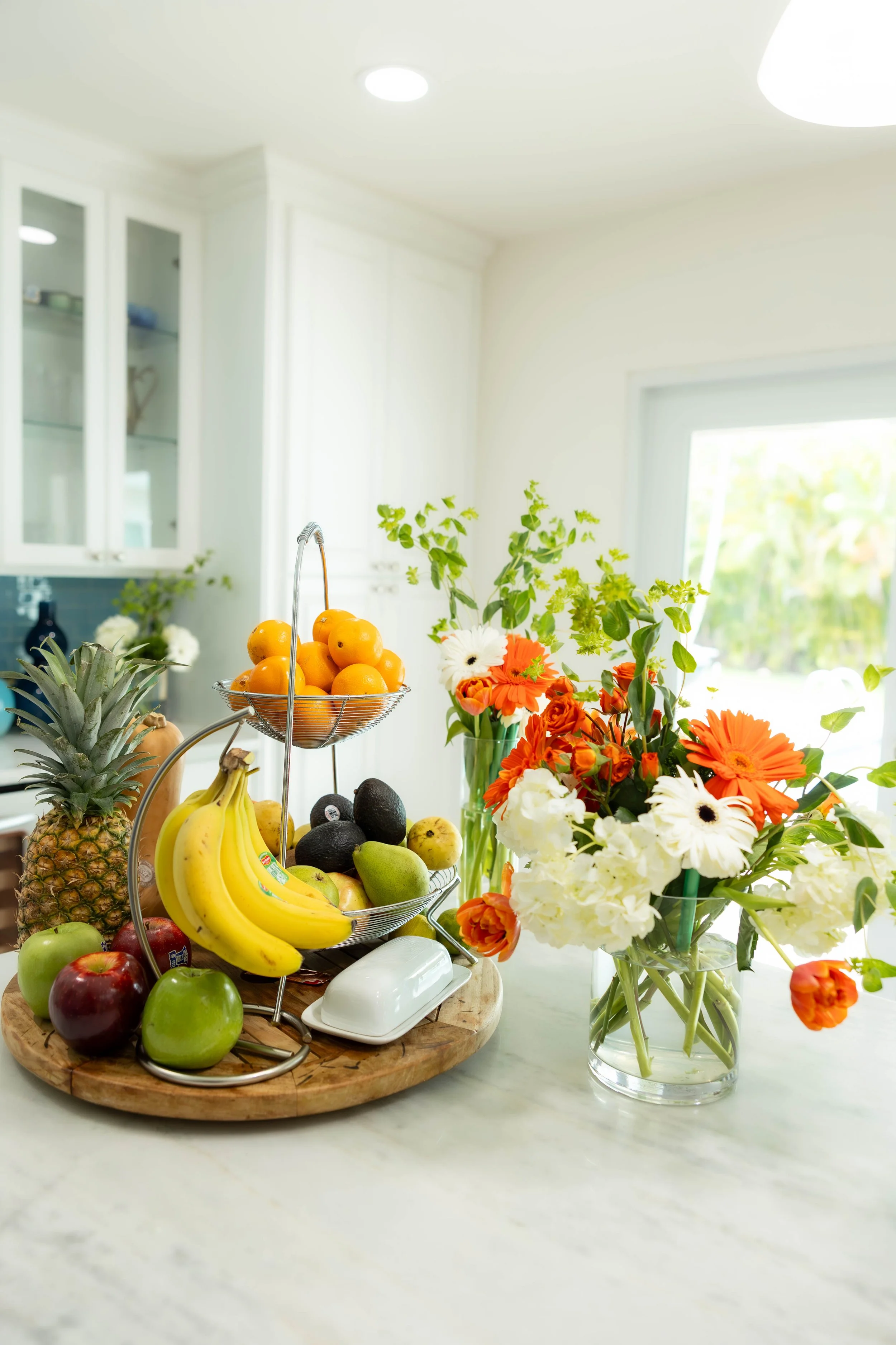 Arrangement of fresh fruit including pineapple, bananas, green and red apples, avocados, and oranges displayed on a wooden tray. Bright flowers in a glass vase beside the tray, with a background of a bright kitchen space.