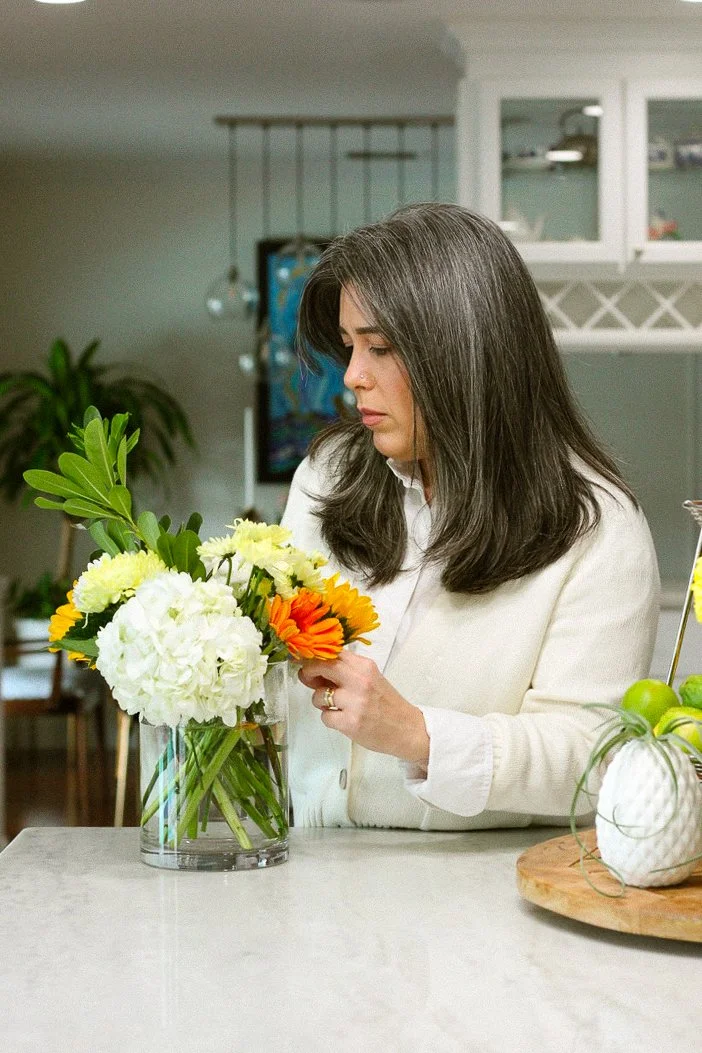 A woman with long dark hair arranging a bouquet of white, yellow, and orange flowers in a clear glass vase on a kitchen countertop.