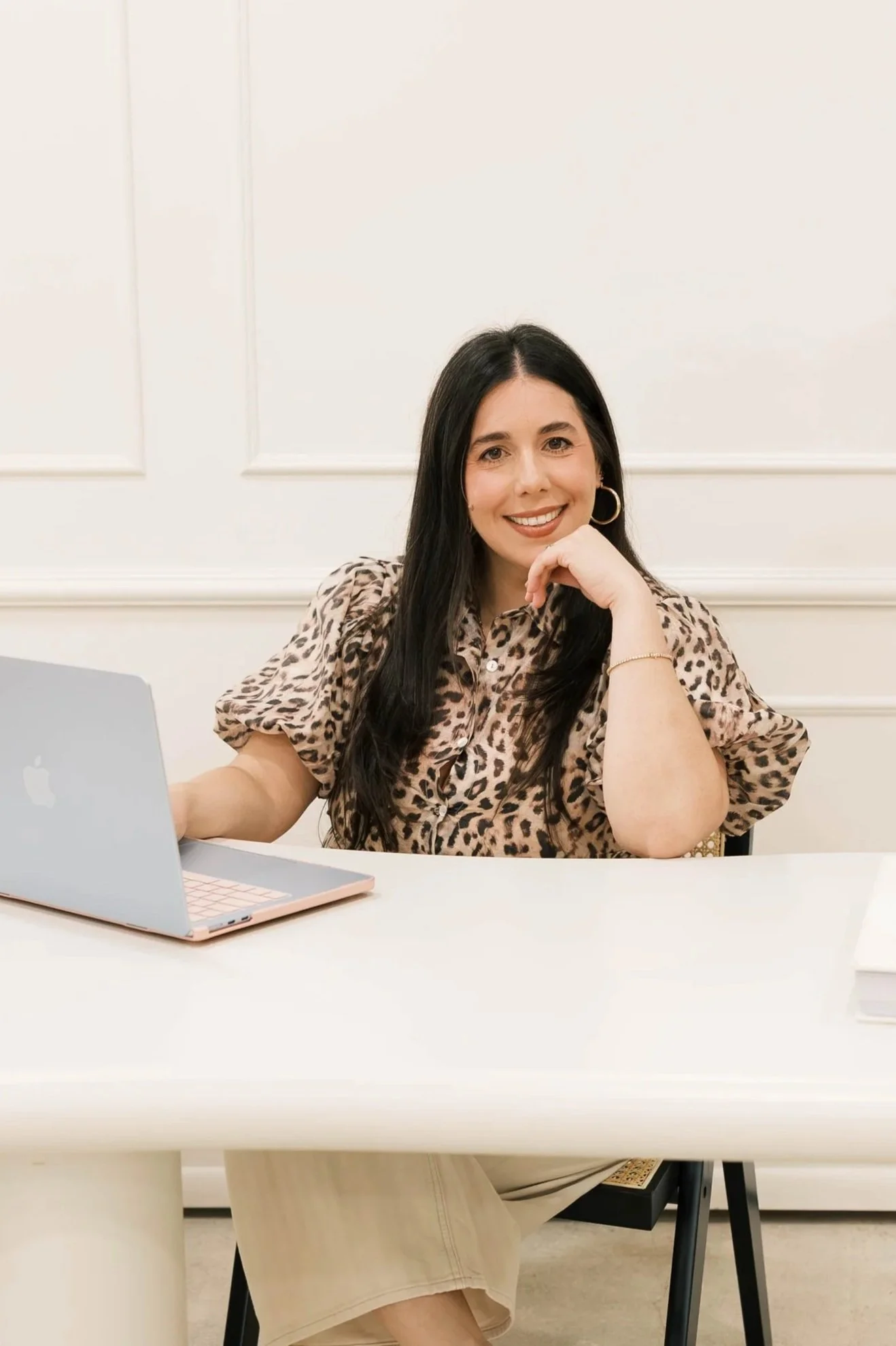 A woman sitting at a white table with a laptop, smiling, wearing a leopard print blouse, with white panel walls in the background.