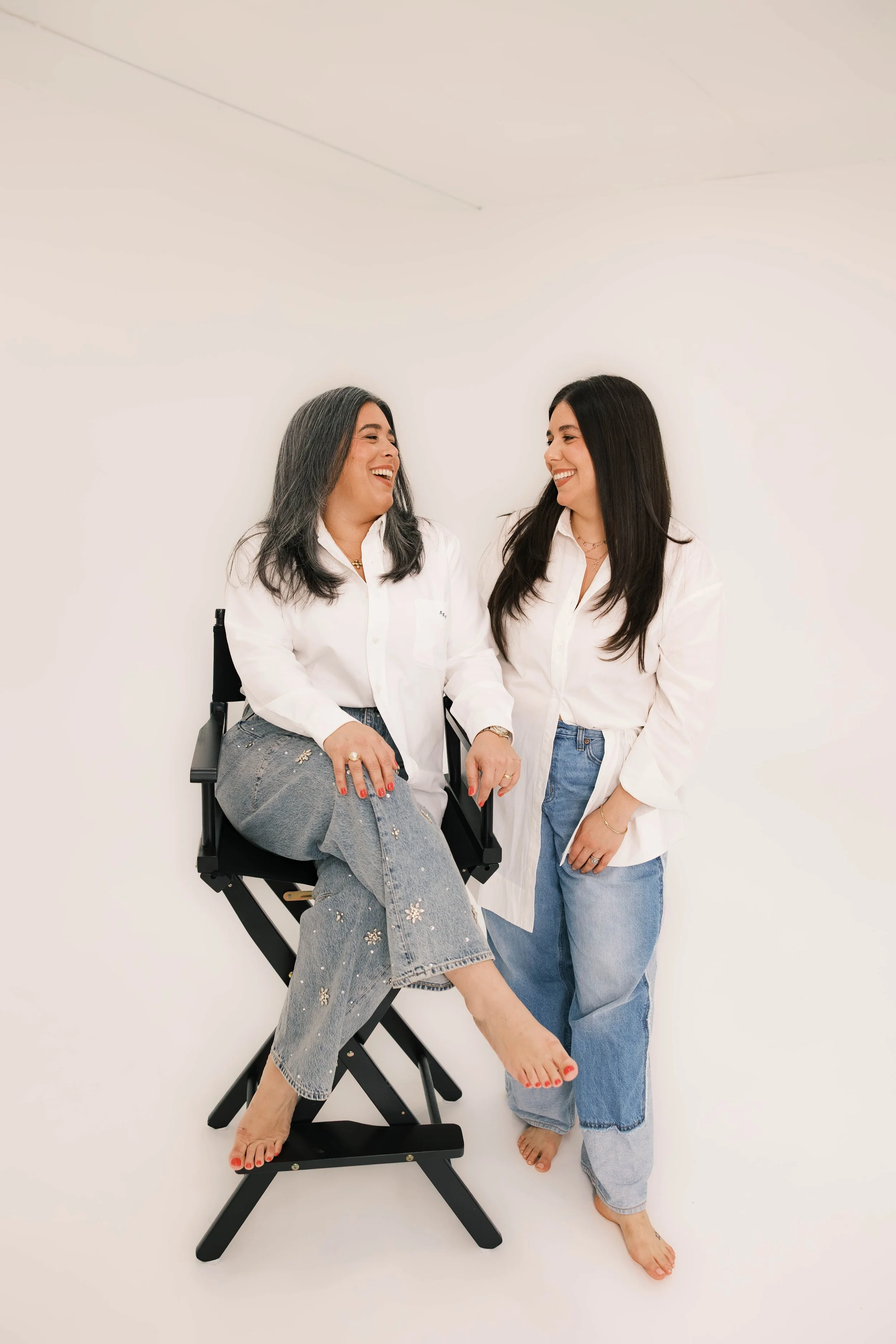Two women with dark hair laughing and smiling, wearing white shirts and jeans, one sitting on a director's chair and the other standing, against a plain white background.