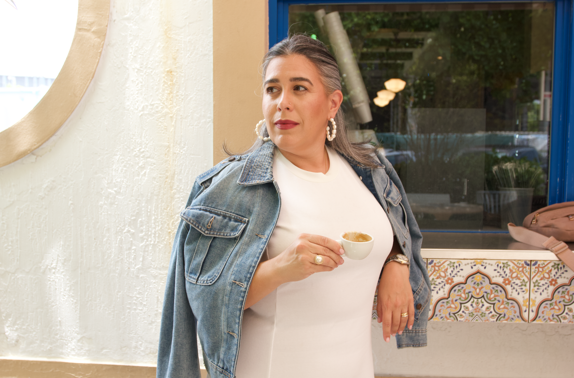 A woman with gray hair wearing a denim jacket and white top, holding a small white cup, standing inside a cafe with colorful tiled counter and large window.