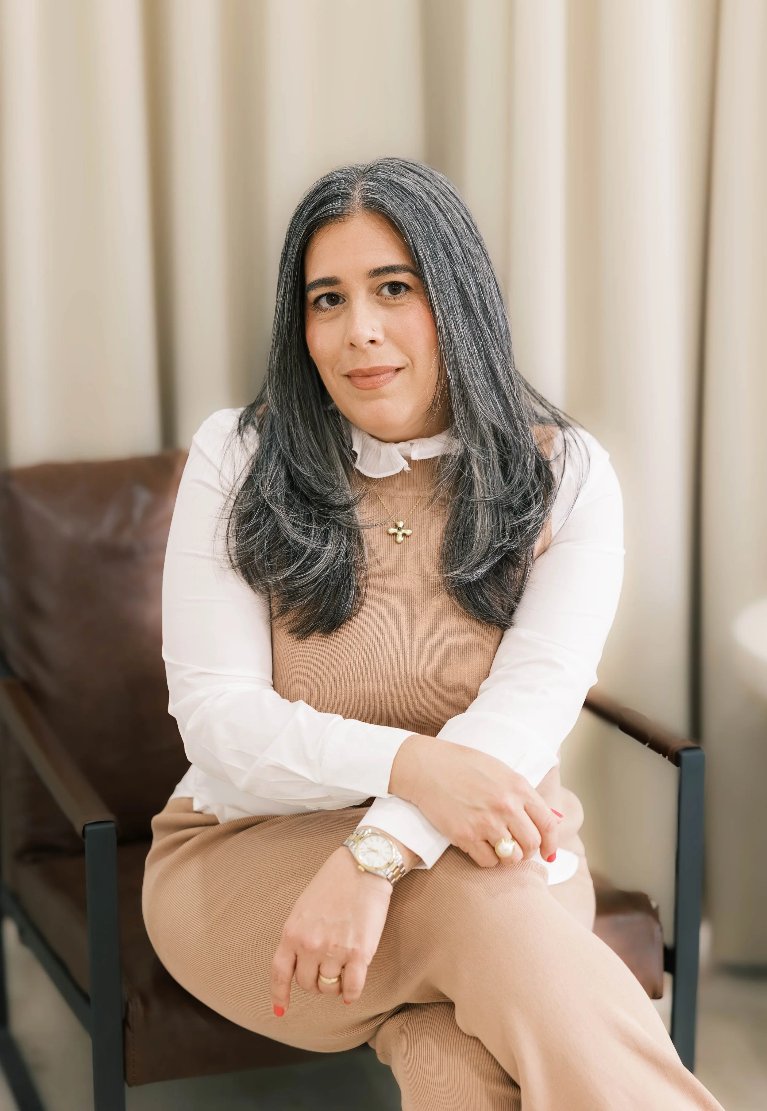 A woman with long, dark hair with gray streaks, sitting on a brown chair in front of beige curtains, wearing a white blouse with a ruffled collar underneath a beige sleeveless top, beige pants, gold jewelry, and a wristwatch.