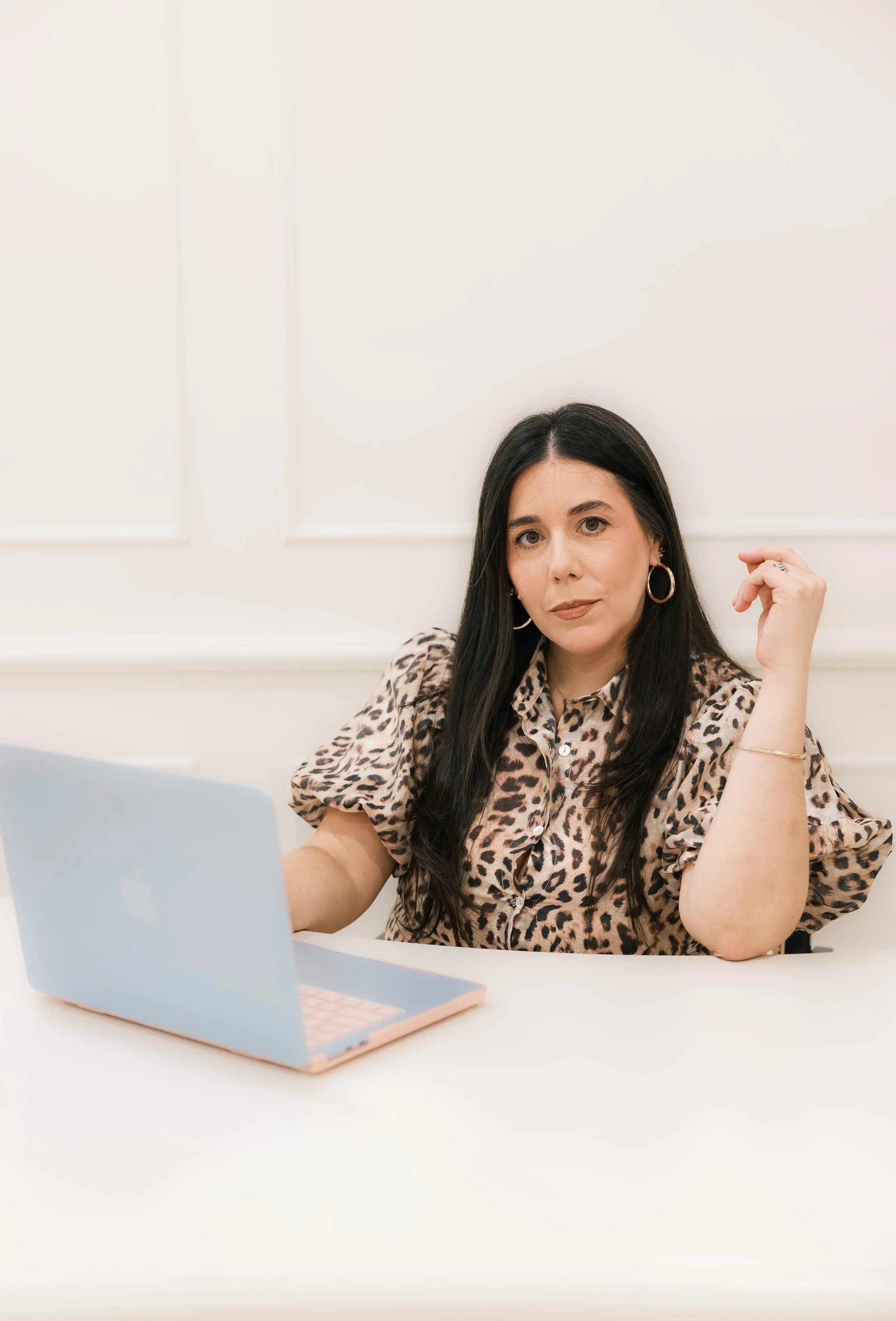 A woman with long dark hair, wearing a leopard print blouse and hoop earrings, sitting at a white desk with an open pastel pink and blue laptop in front of her, in a bright room with white walls.