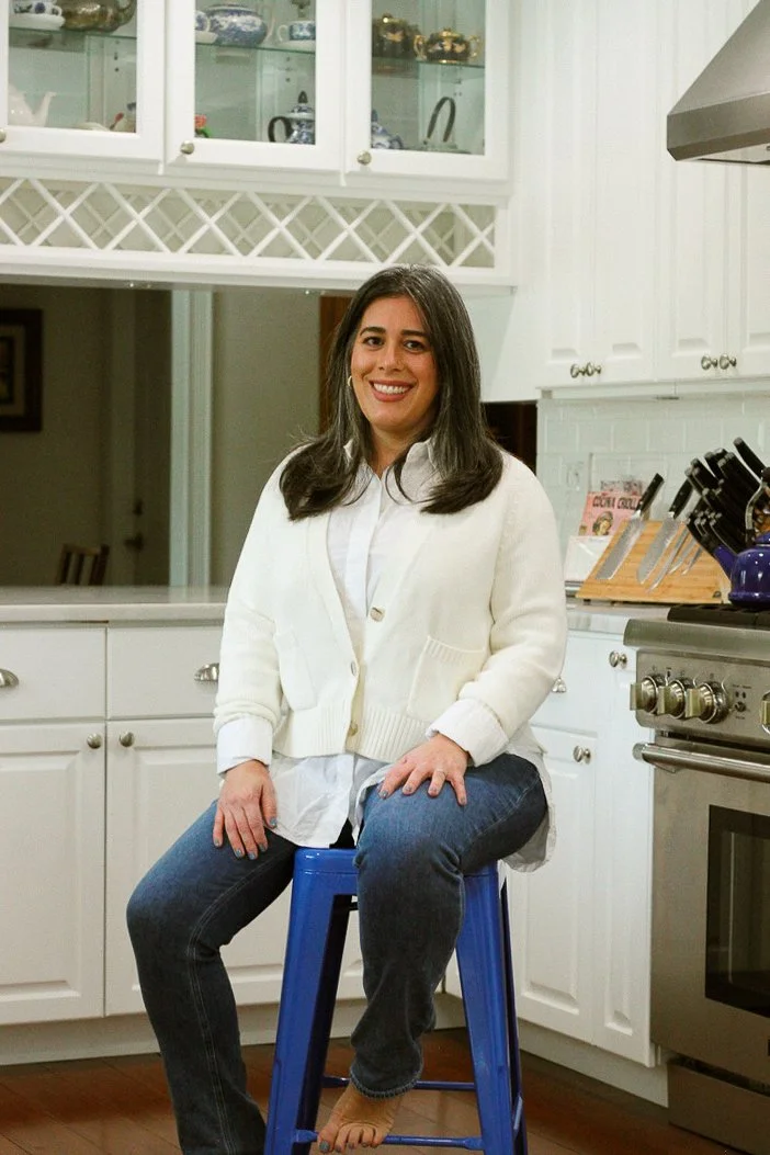A woman with long dark hair sits on a blue stool in a kitchen, smiling at the camera. She is wearing a white sweater and dark jeans, with a white shirt underneath. The kitchen features white cabinets, a stove, and various kitchen items on the counter.