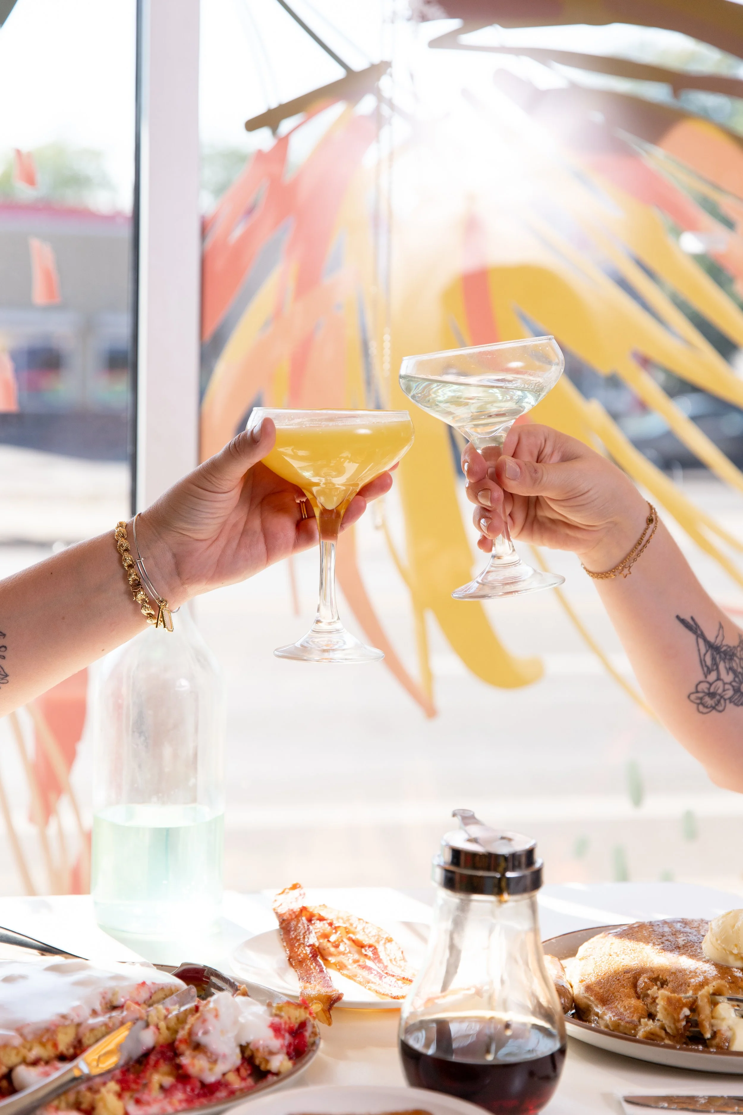 Two people clinking glasses of cocktails at a breakfast table with food, a bottle, and a colorful umbrella in the background.