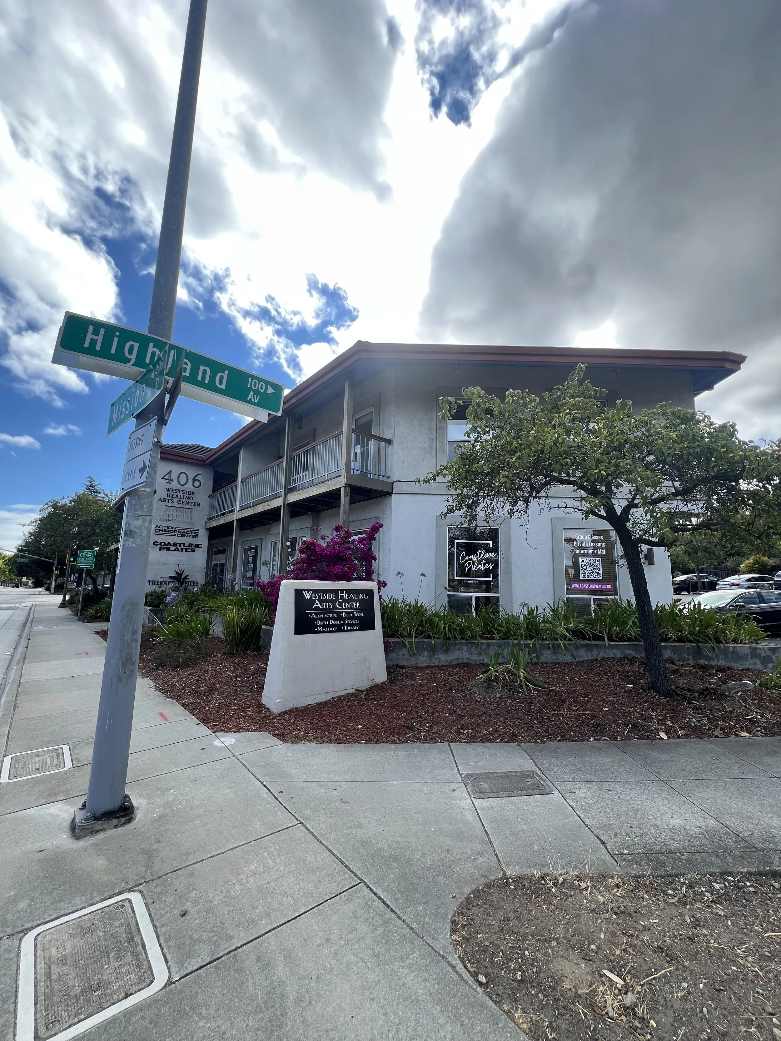A street corner with a building housing the Westside Healing Arts Center, with signboard, pink flowering shrub, tree, and a crosswalk.