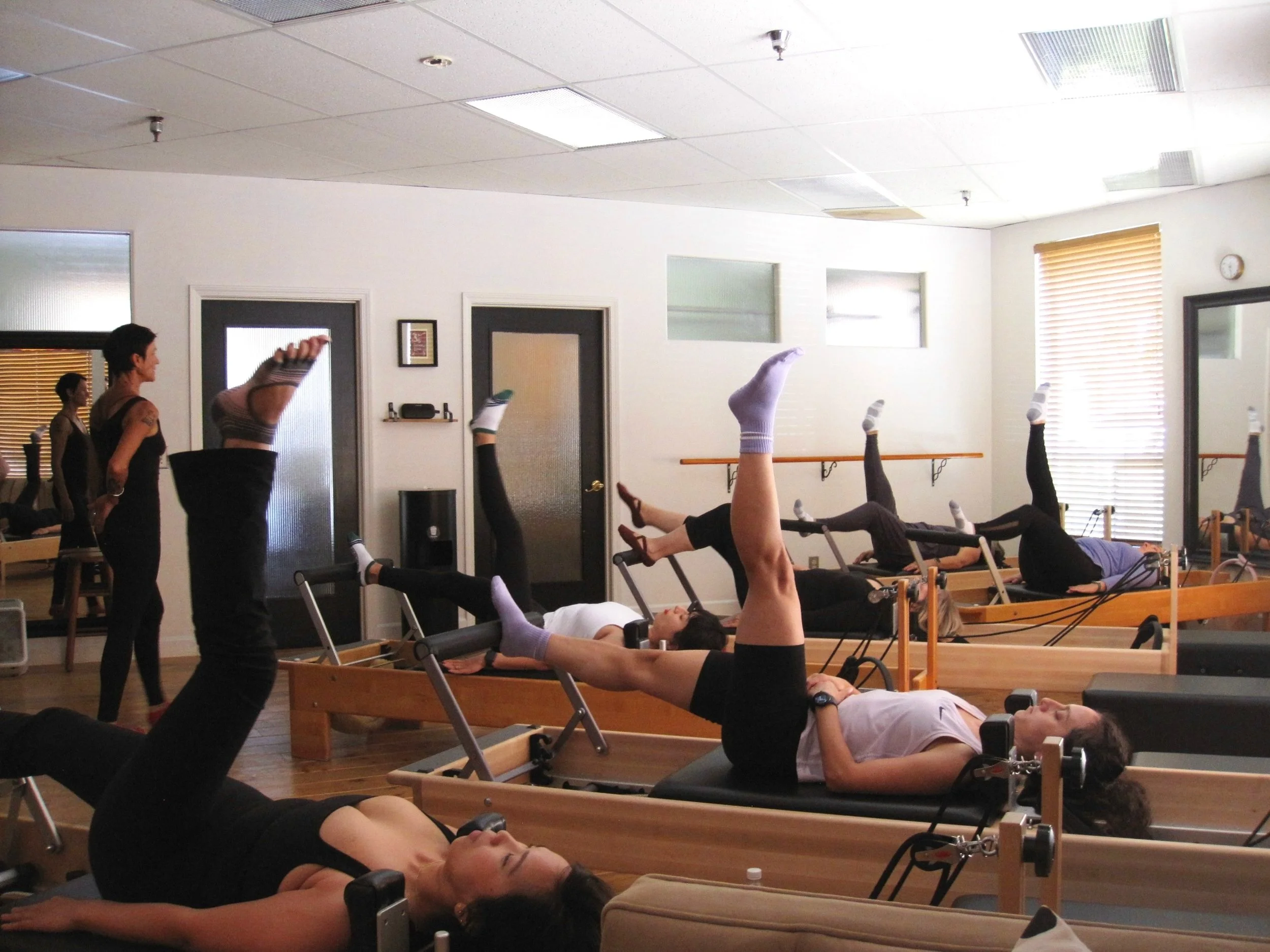 People participating in a Pilates class in a studio, lying on reformer machines with one leg raised.