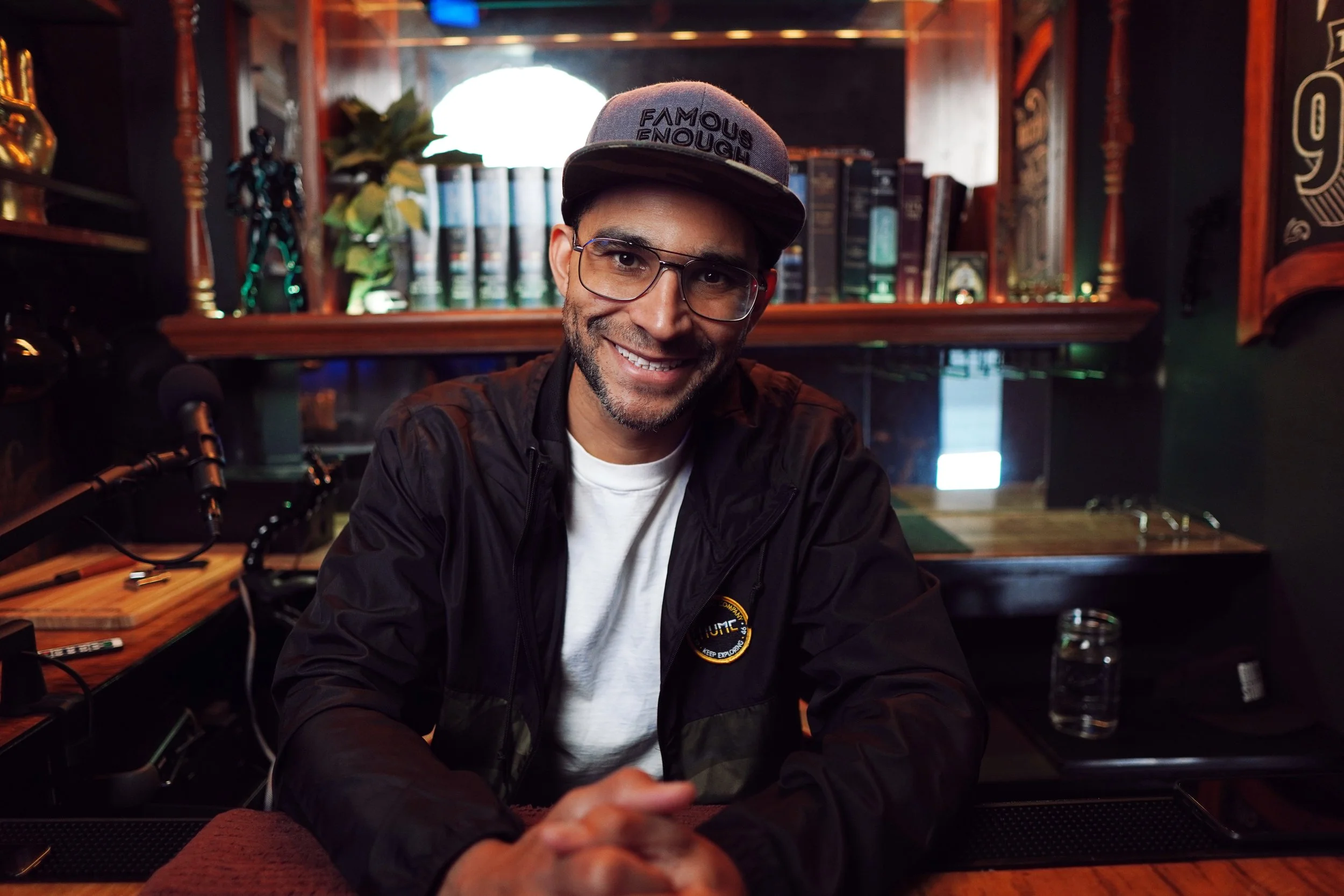 A man with glasses smiling, wearing a gray cap with 'FAMOUS ENOUGH' written on it, sitting at a bar with bookshelves and plants in the background.
