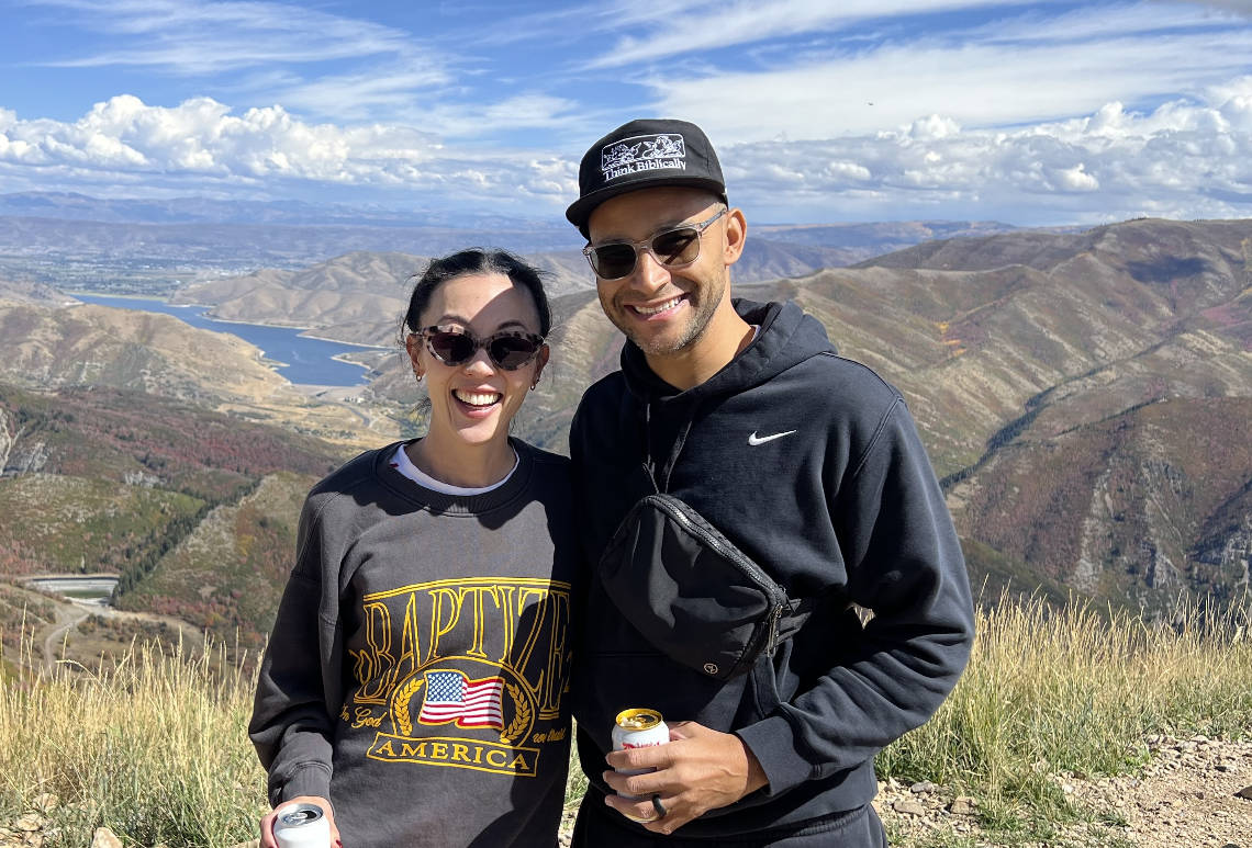 A smiling couple with sunglasses on a mountain overlooking a reservoir and colorful landscape, enjoying a sunny day.
