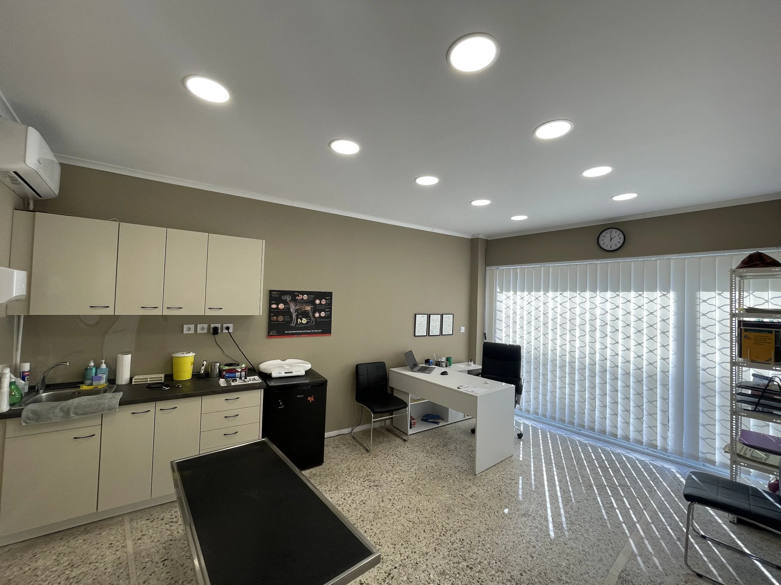 Medical office room with beige cabinets, medical equipment, a desk with a chair, and medical posters. Large window with vertical blinds, empty chairs, and a metal shelving unit.