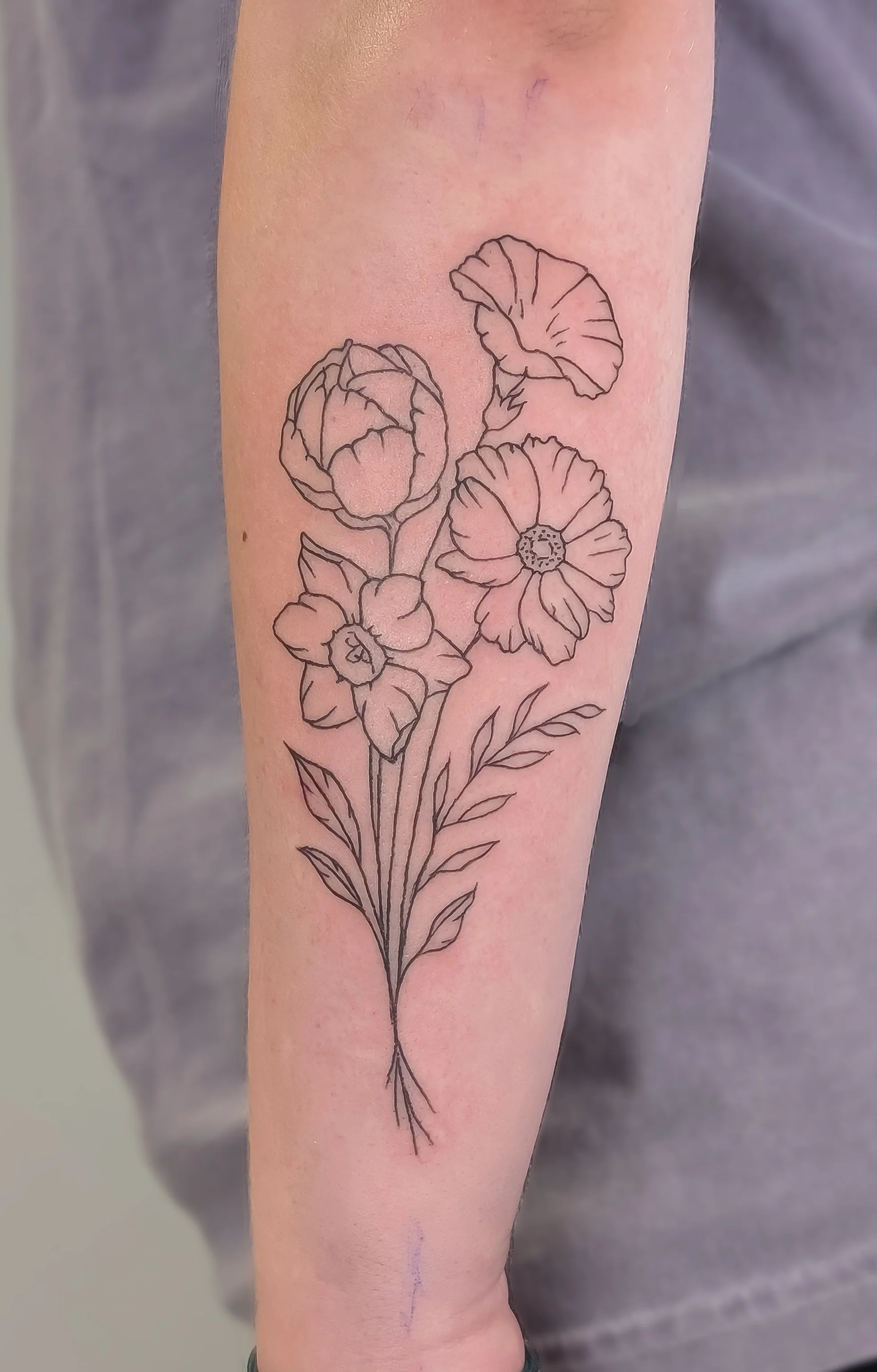 A black outline tattoo of a bouquet of flowers on a person's forearm, featuring different types of blooming flowers and leaves.