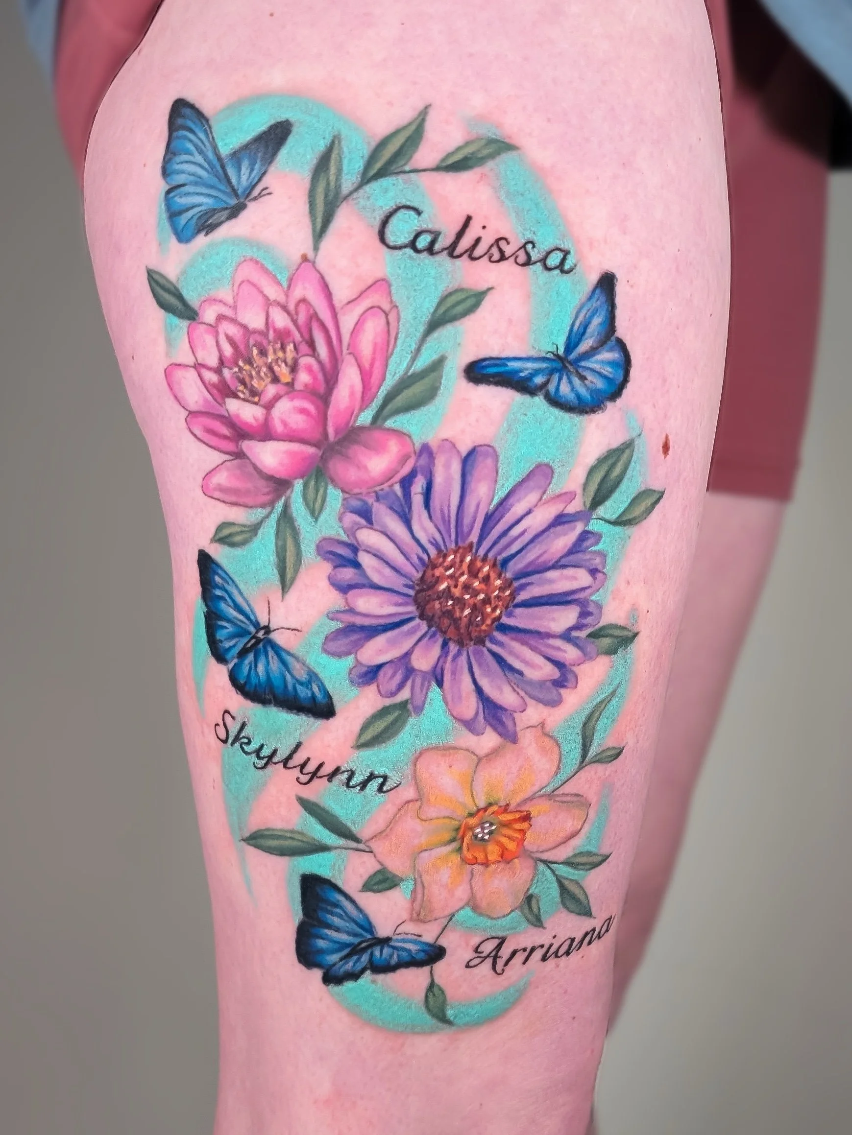 A tattoo of pink and purple flowers, green leaves, and blue butterflies on a person's skin. The tattoo includes the names 'Calissa,' 'Skylan,' and 'Arriano' written in cursive.