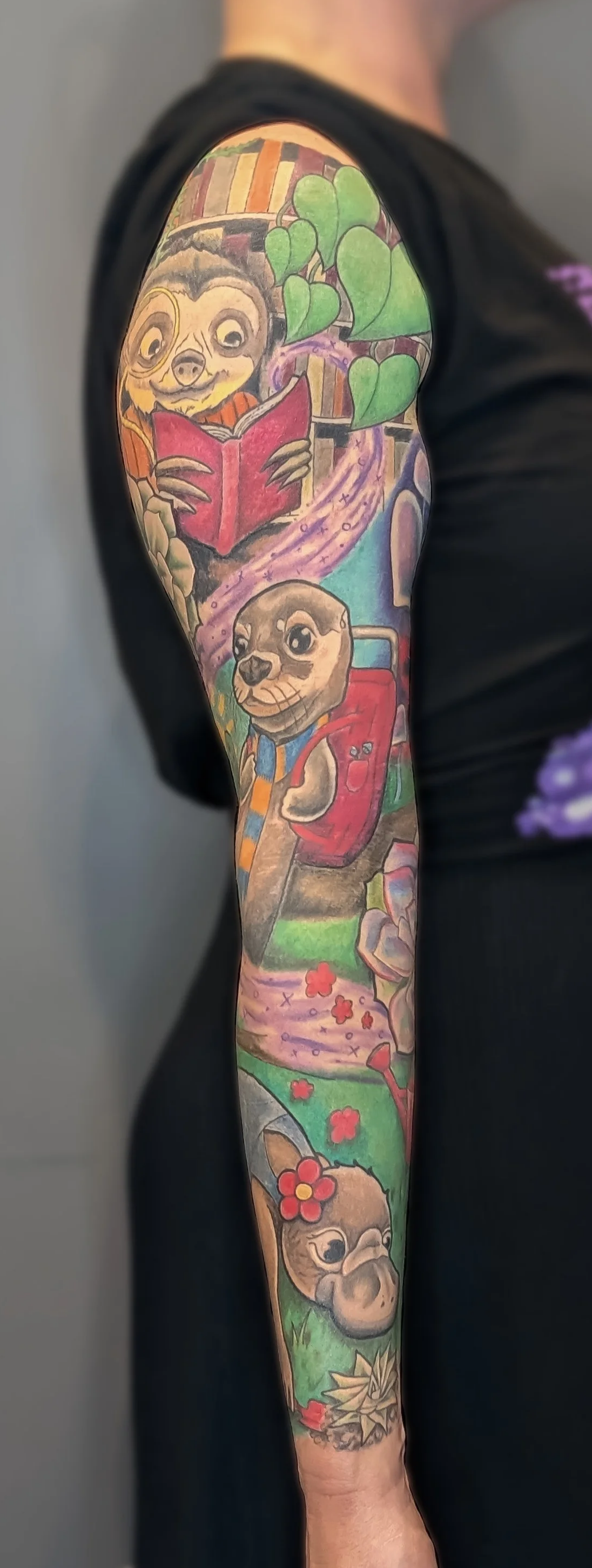 Colorful arm tattoo featuring cartoon animals, a book, a rainbow path, and greenery.