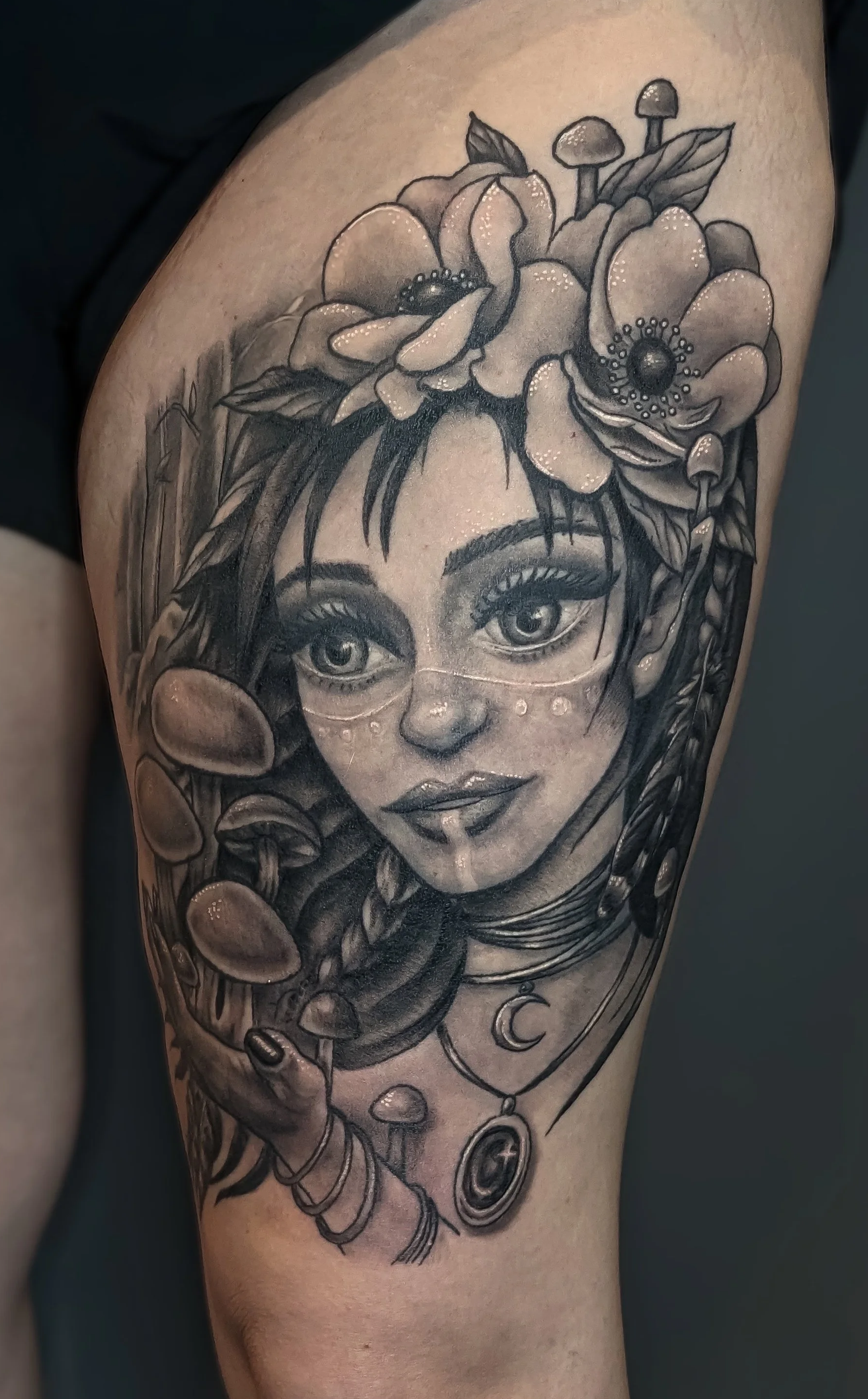 Black and gray tattoo of a woman's face with flowers and jewelry on her forehead and around her neck.