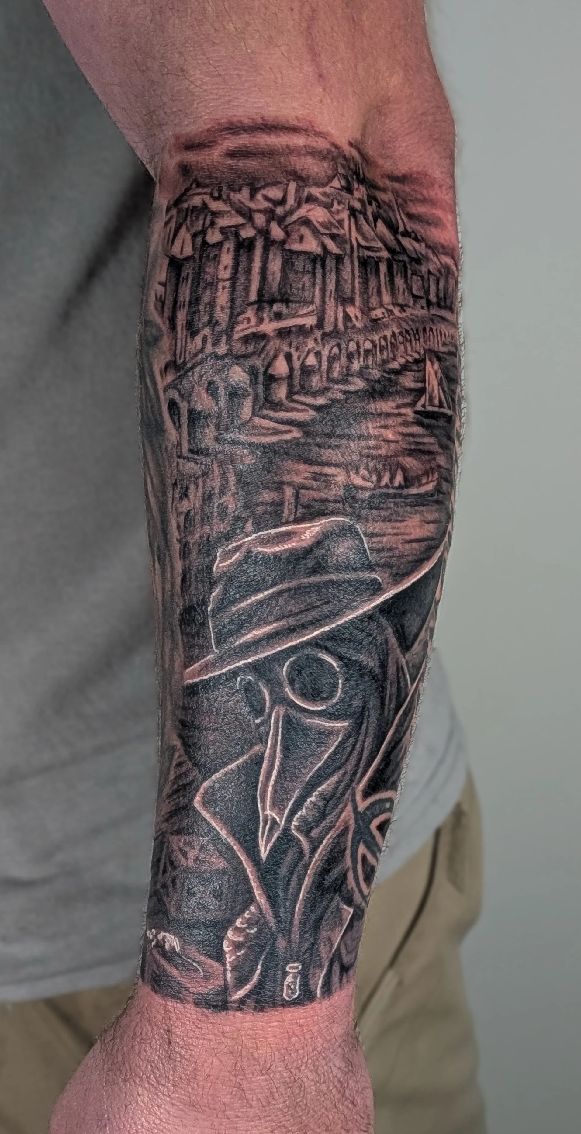 Black and gray tattoo on a person's forearm featuring a detailed seaside scene with houses, boats, and a person holding a fishing rod.