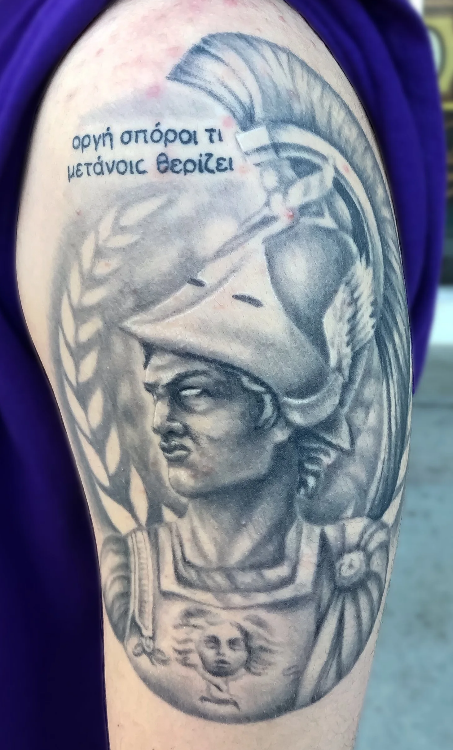 Tattoo of a Greek mythological figure on the arm, depicting a helmeted woman with a serious expression, surrounded by decorative elements and Greek text.