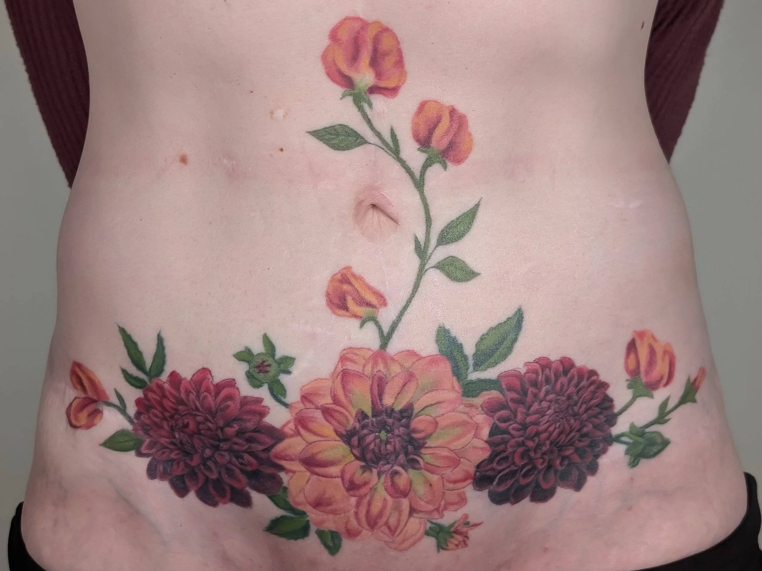 A tattoo of a floral design with large orange, purple, and pink flowers with green leaves on a person's stomach.