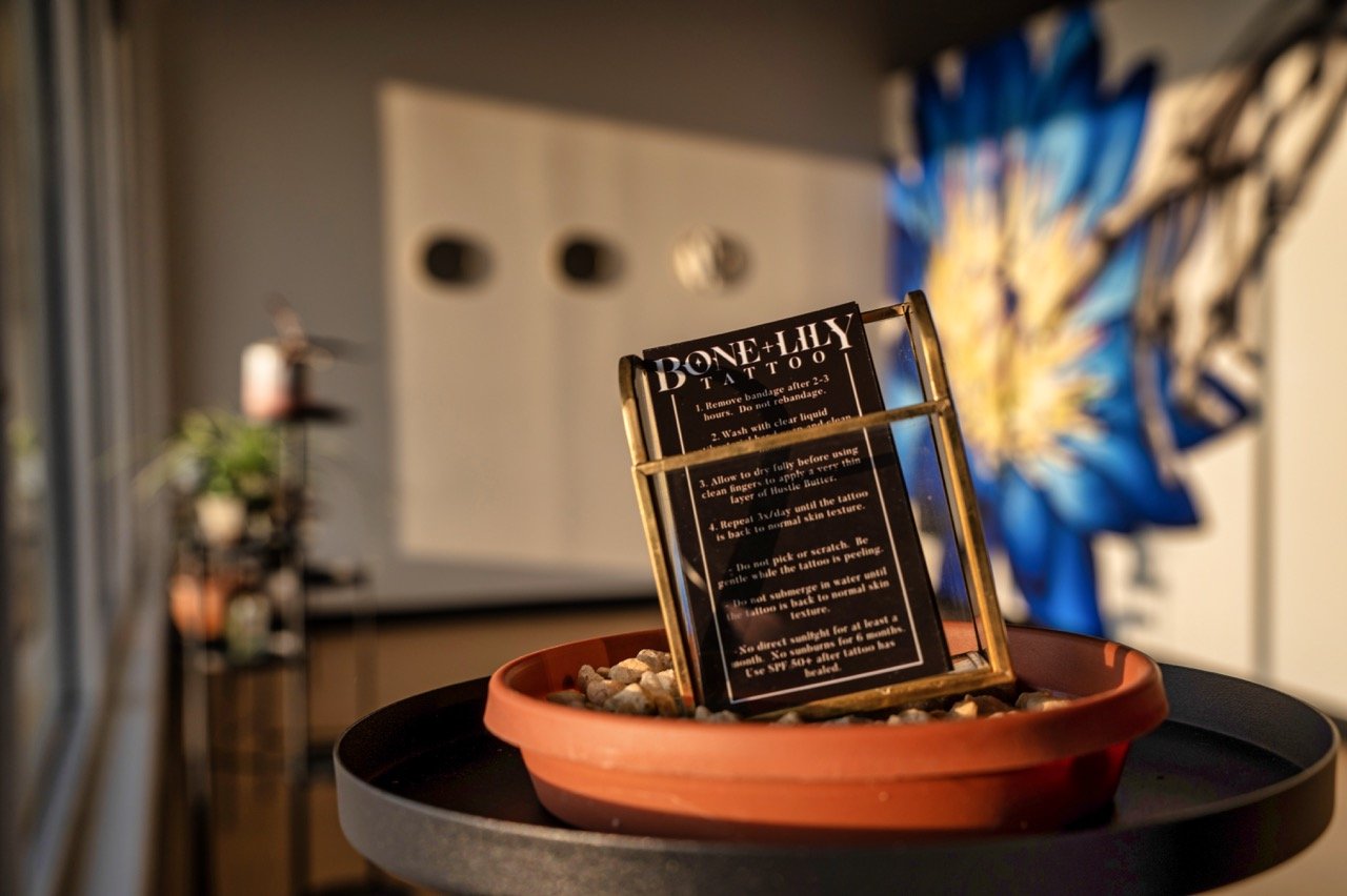 Tattoo aftercare instructions displayed on a black sign inside a transparent holder, placed on a round surface with a small brown container filled with small rocks, in a tattoo studio with artwork on the wall and a blurred background.