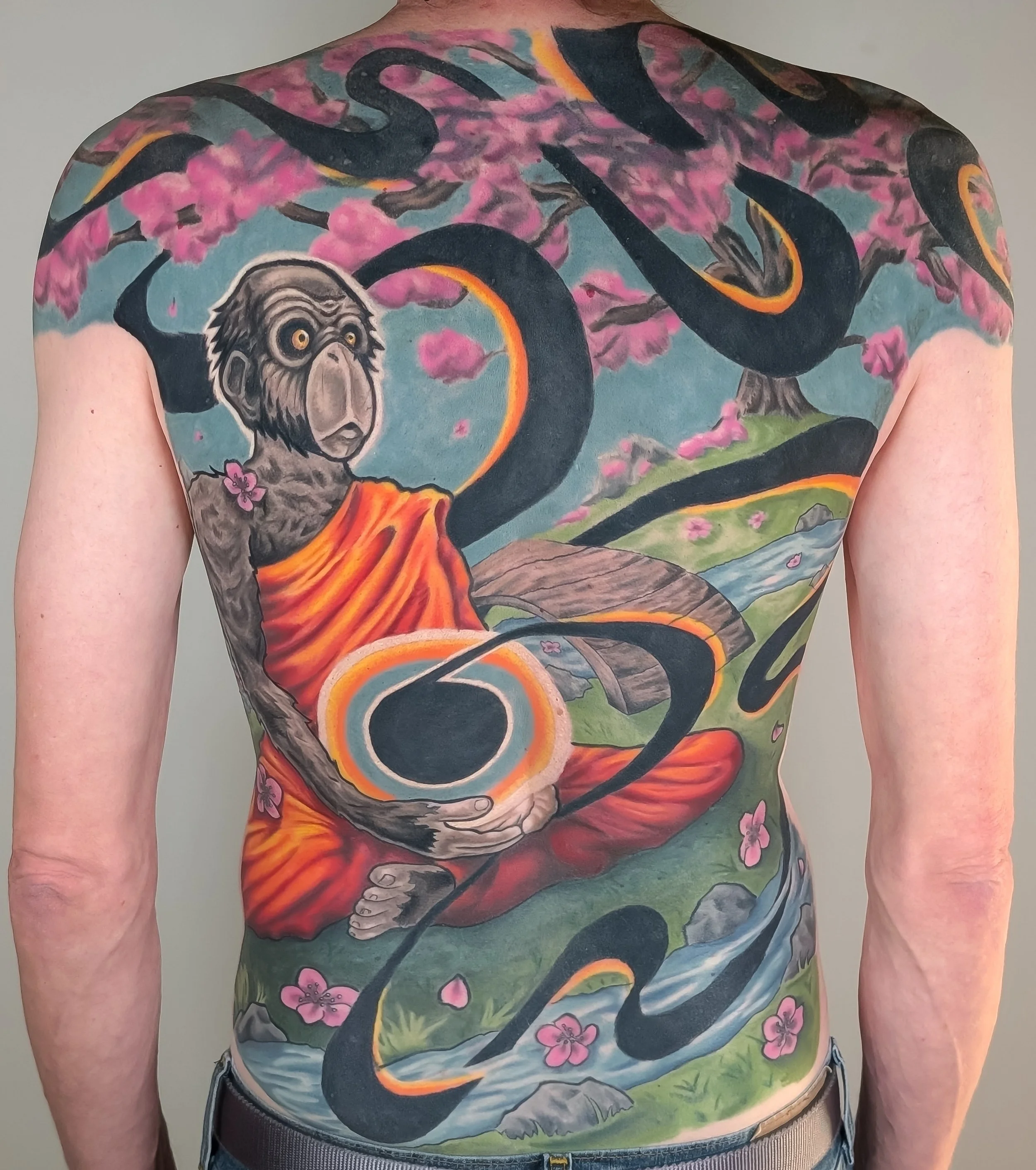 A person with a rainbow-colored tattoo of a dragon and a monkey on their back, featuring pink blossoms and a bright blue sky.