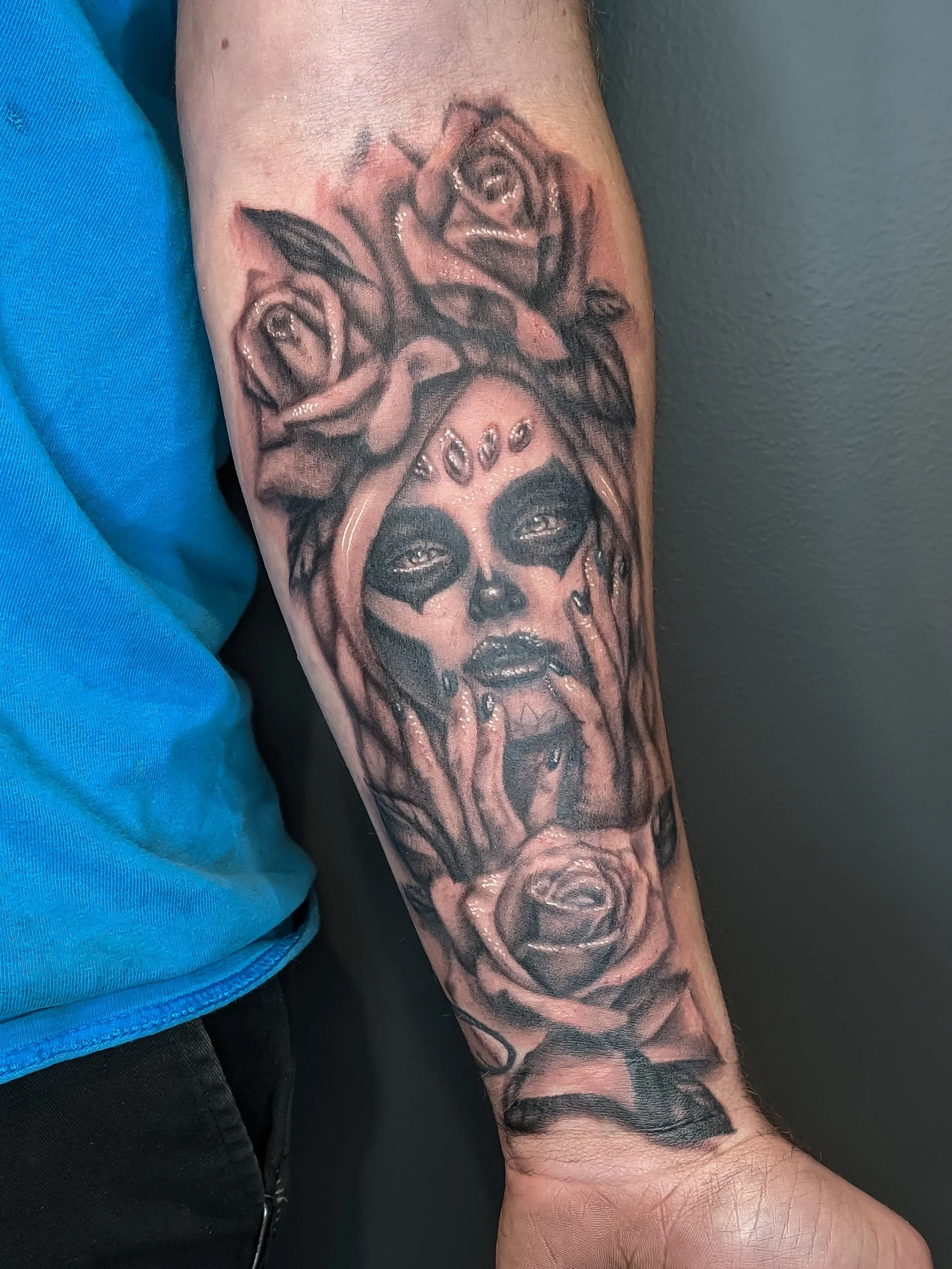 Close-up of a person's forearm tattoo featuring a face with dark makeup, surrounded by roses and other floral designs.