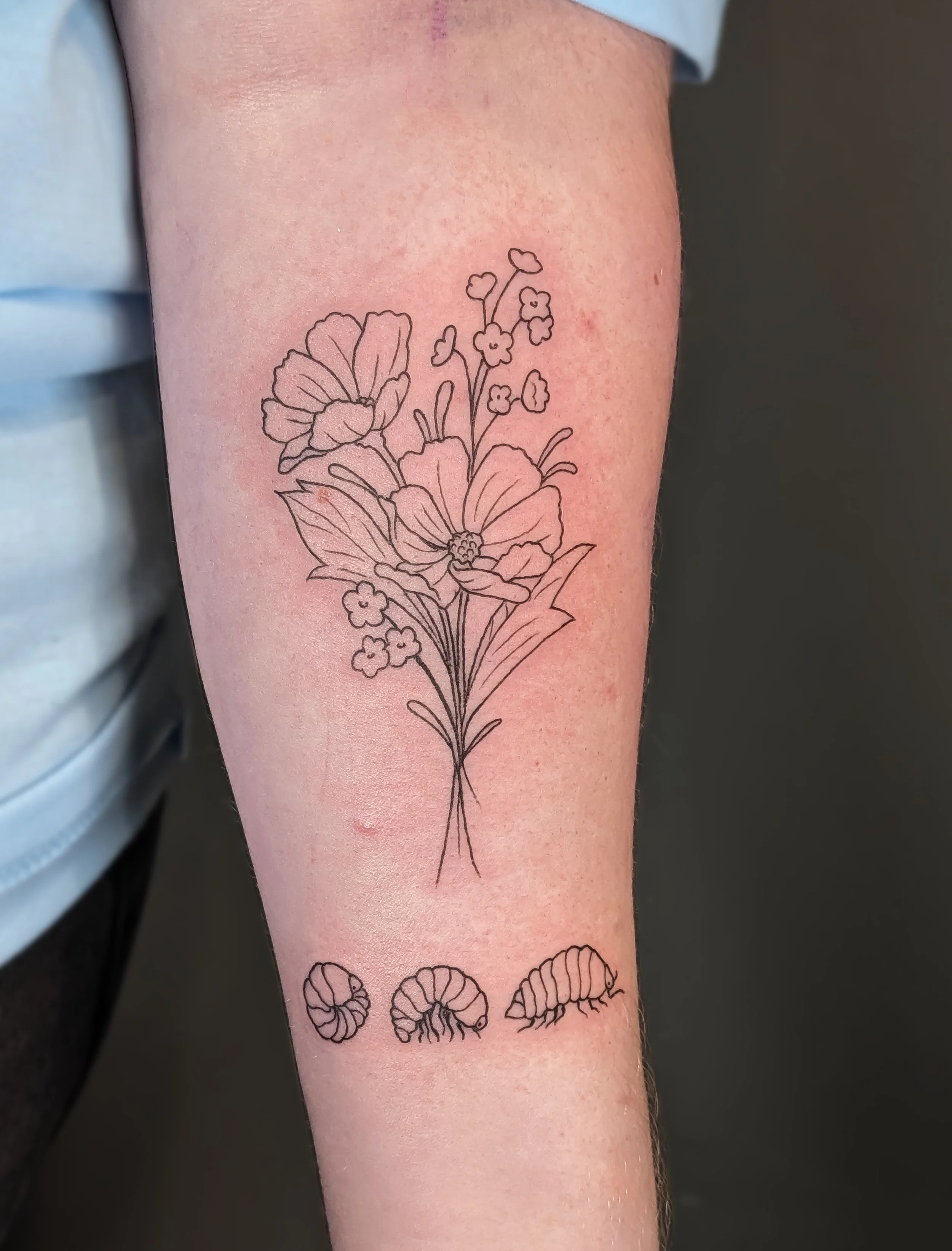 Tattoo of a floral bouquet with large flowers and smaller blossoms on upper arm, with a row of curled shell designs below.